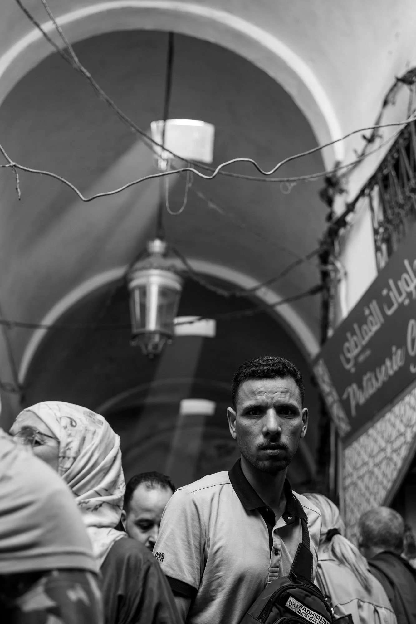  Young man looking straight at the camera in a crowded vaulted souk, medina of Tunis. Black-and-white street photograph by Chris Perona. 