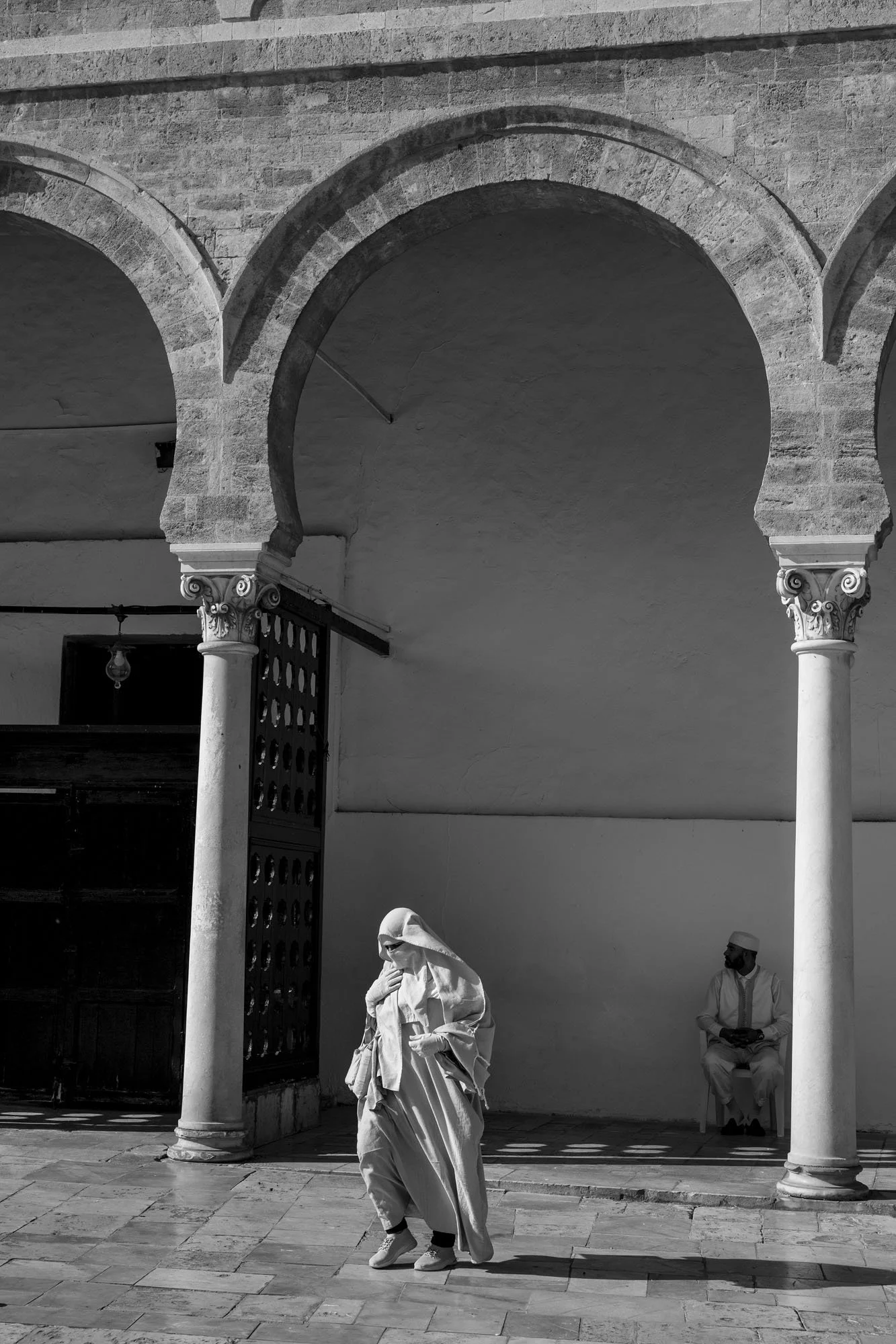  Man in a traditional hooded robe crossing an arcaded courtyard, medina of Tunis. Black-and-white street photograph by Chris Perona. 