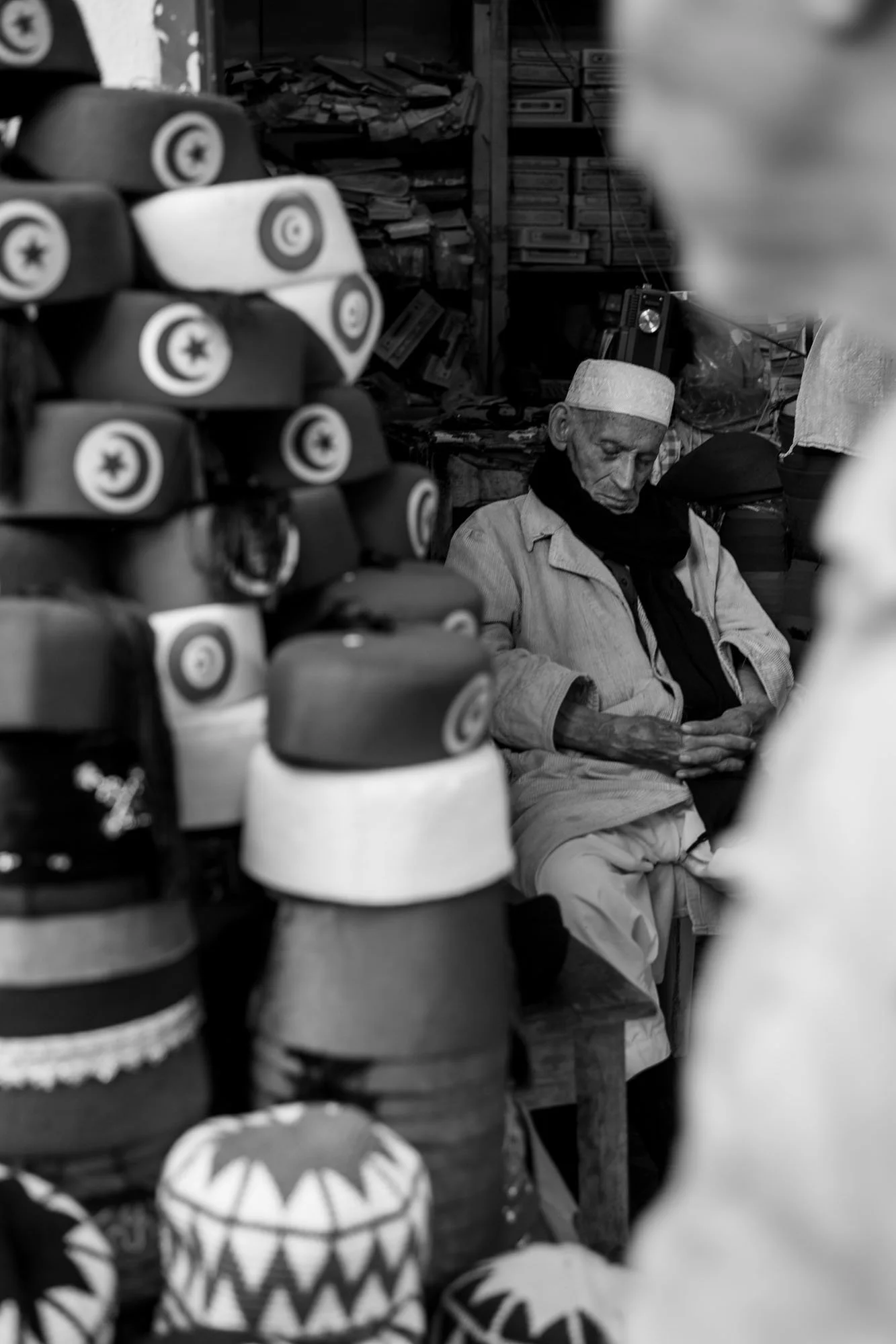  Chechia seller among stacks of traditional caps in the souk, medina of Tunis. Black-and-white street photograph by Chris Perona. 