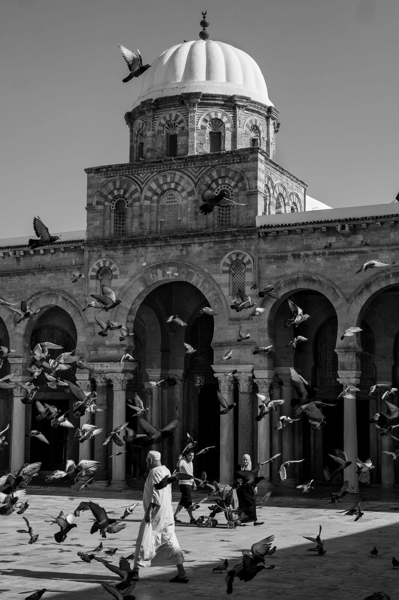  Pigeons taking flight before a domed mosque and its arcades, medina of Tunis. Black-and-white street photograph by Chris Perona. 