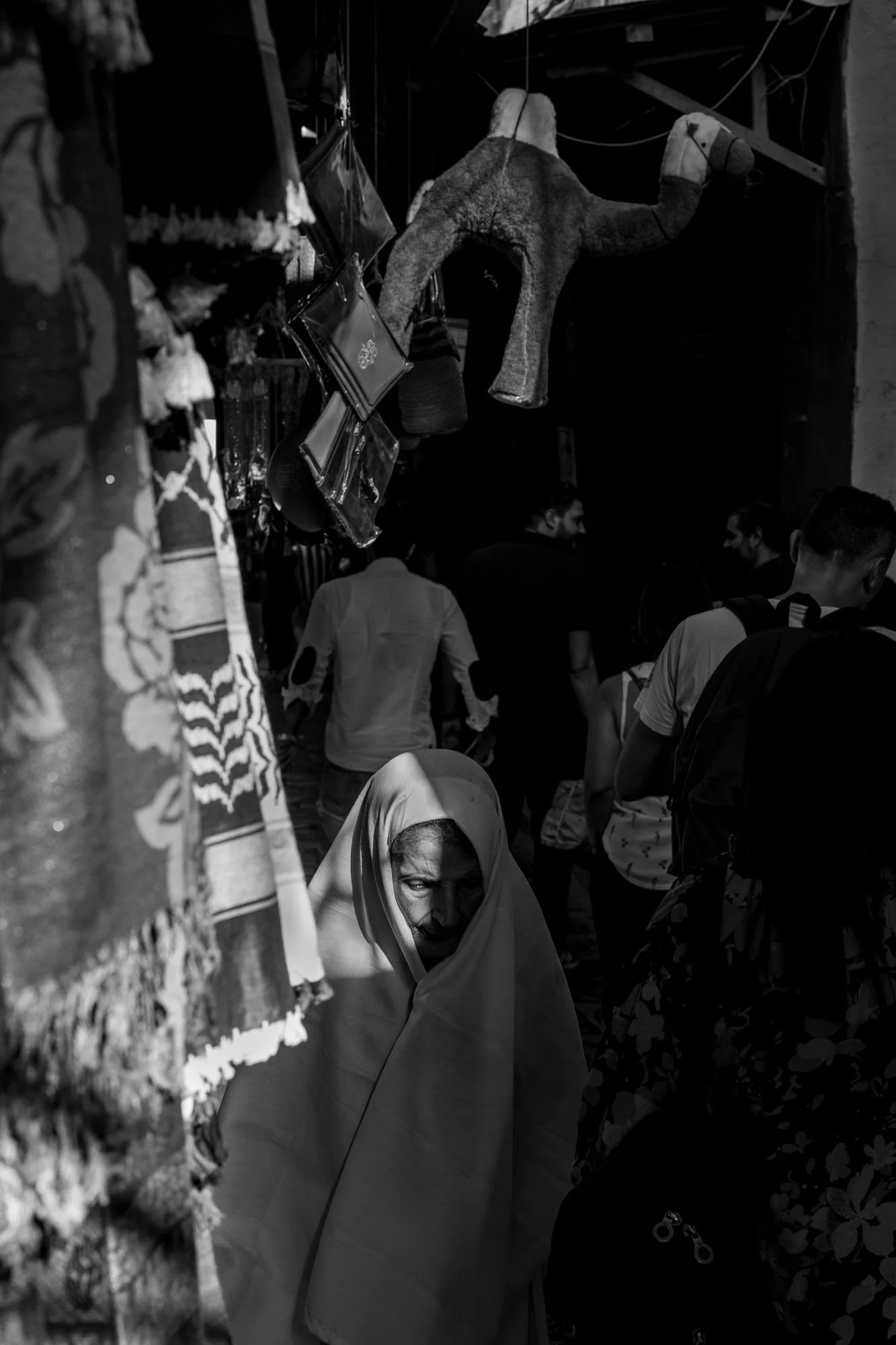  Woman in a headscarf walking through a dim souk hung with textiles, medina of Tunis. Black-and-white street photograph by Chris Perona. 