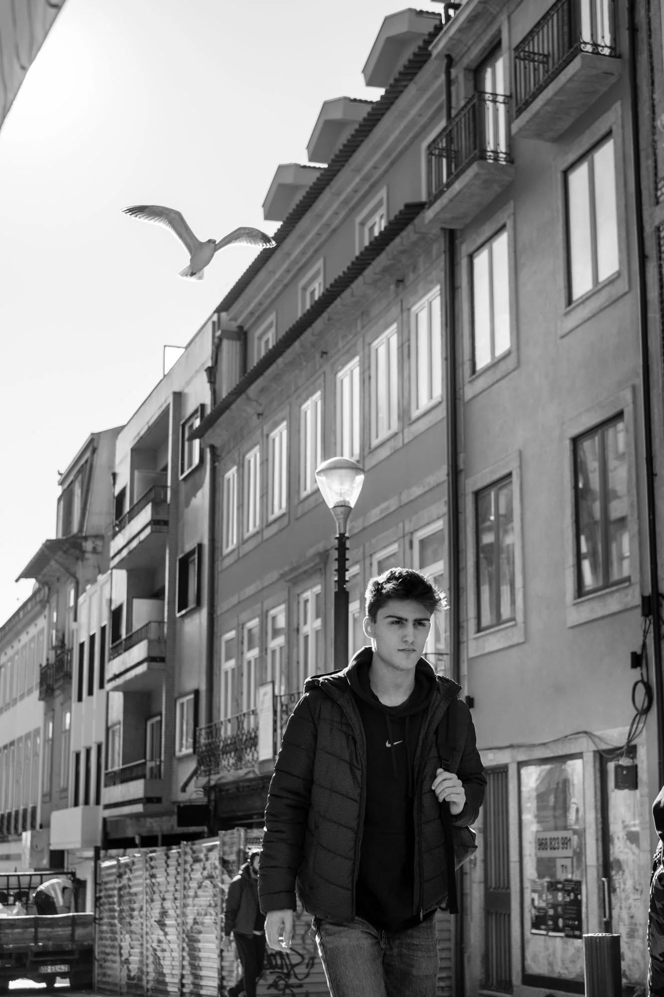  Young man walking along a street of old façades as seagulls fly overhead, Porto. Black-and-white street photograph by Chris Perona. 
