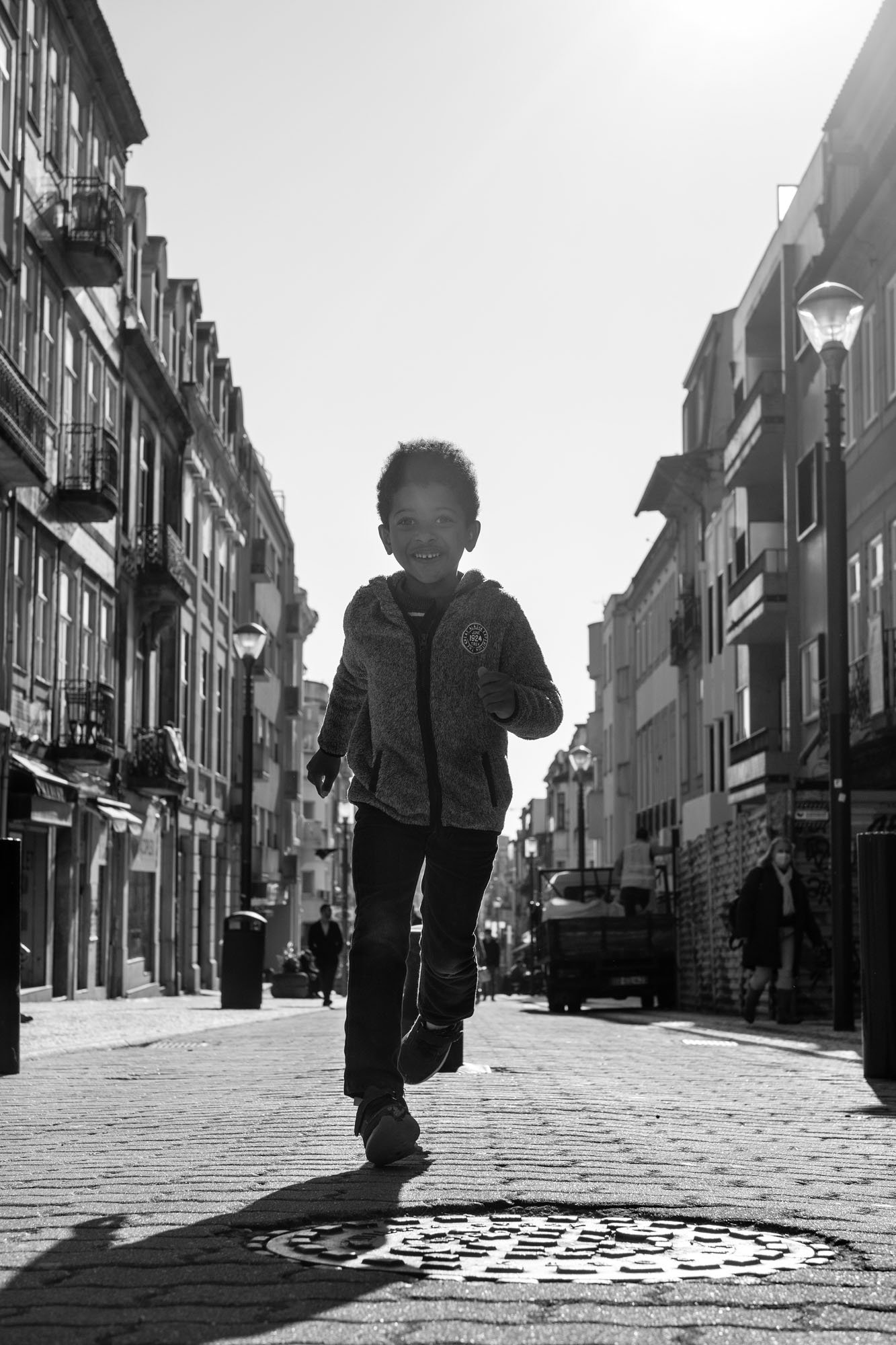 Child running toward the camera on a sunlit cobbled street, Porto. Black-and-white street photograph by Chris Perona. 