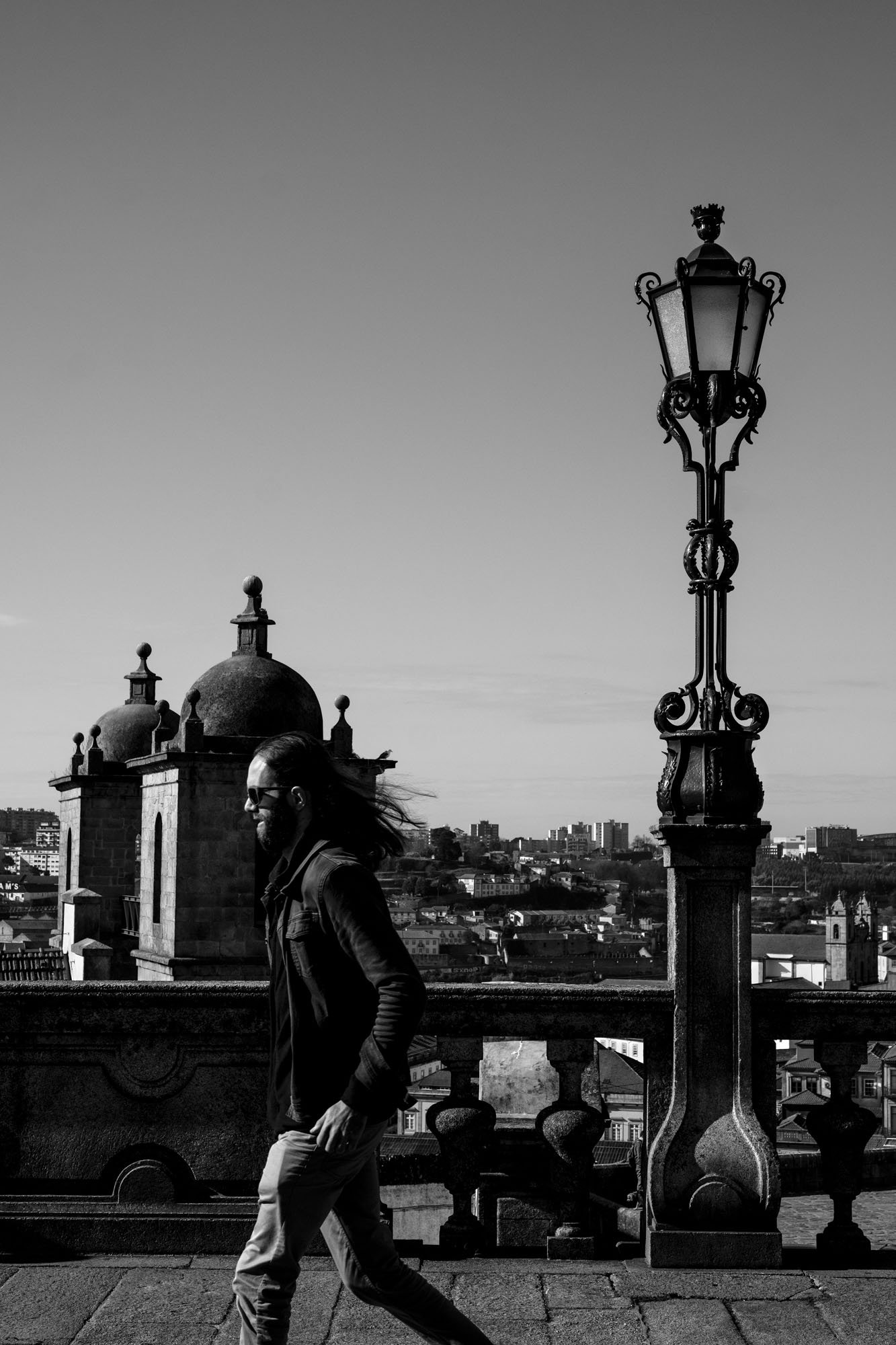  Woman with windswept hair walking past an ornate lamp post above the rooftops, Porto. Black-and-white street photograph by Chris Perona. 