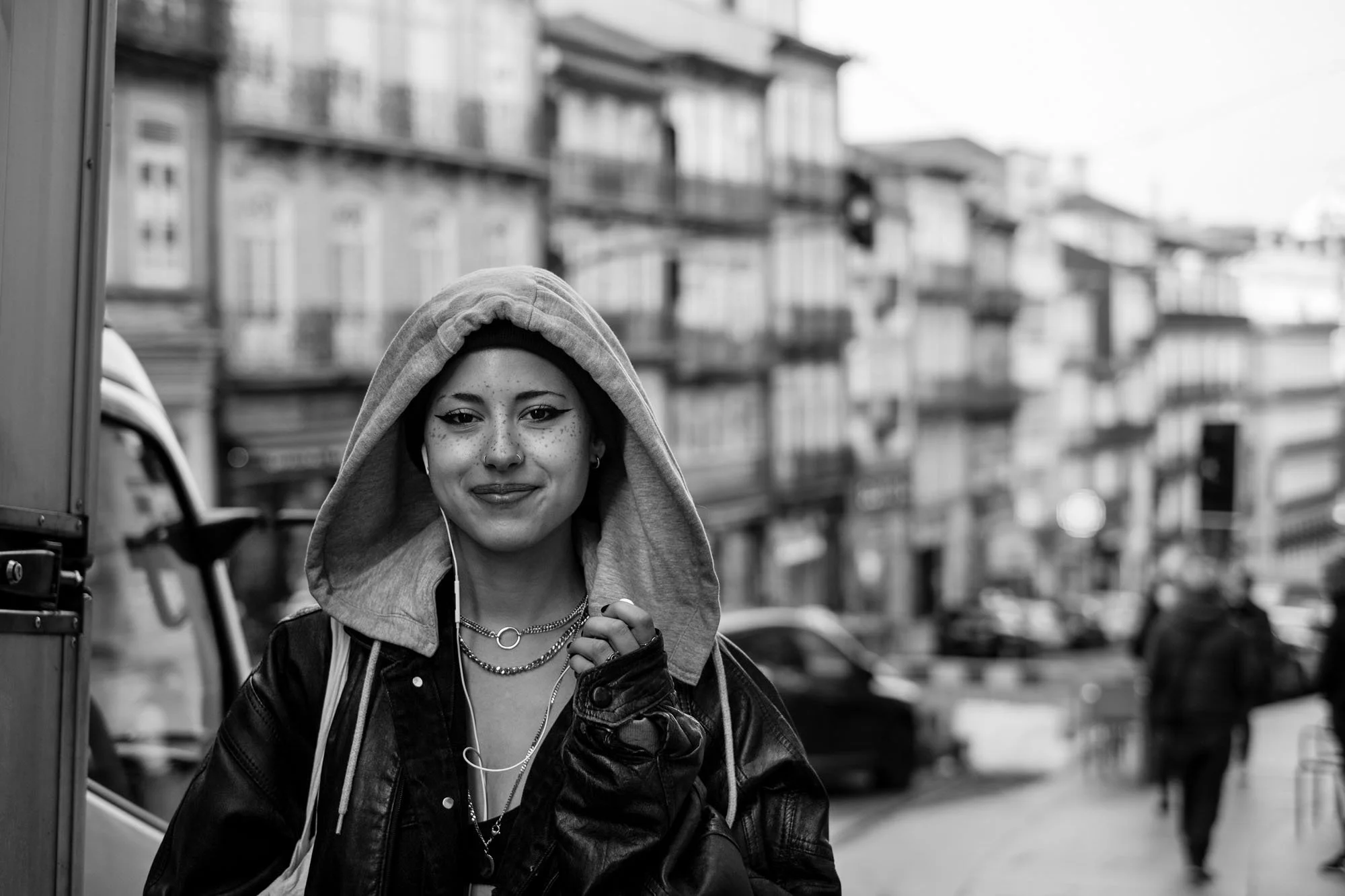  Smiling young woman in a hooded jacket on a busy street, Porto. Black-and-white street portrait by Chris Perona. 