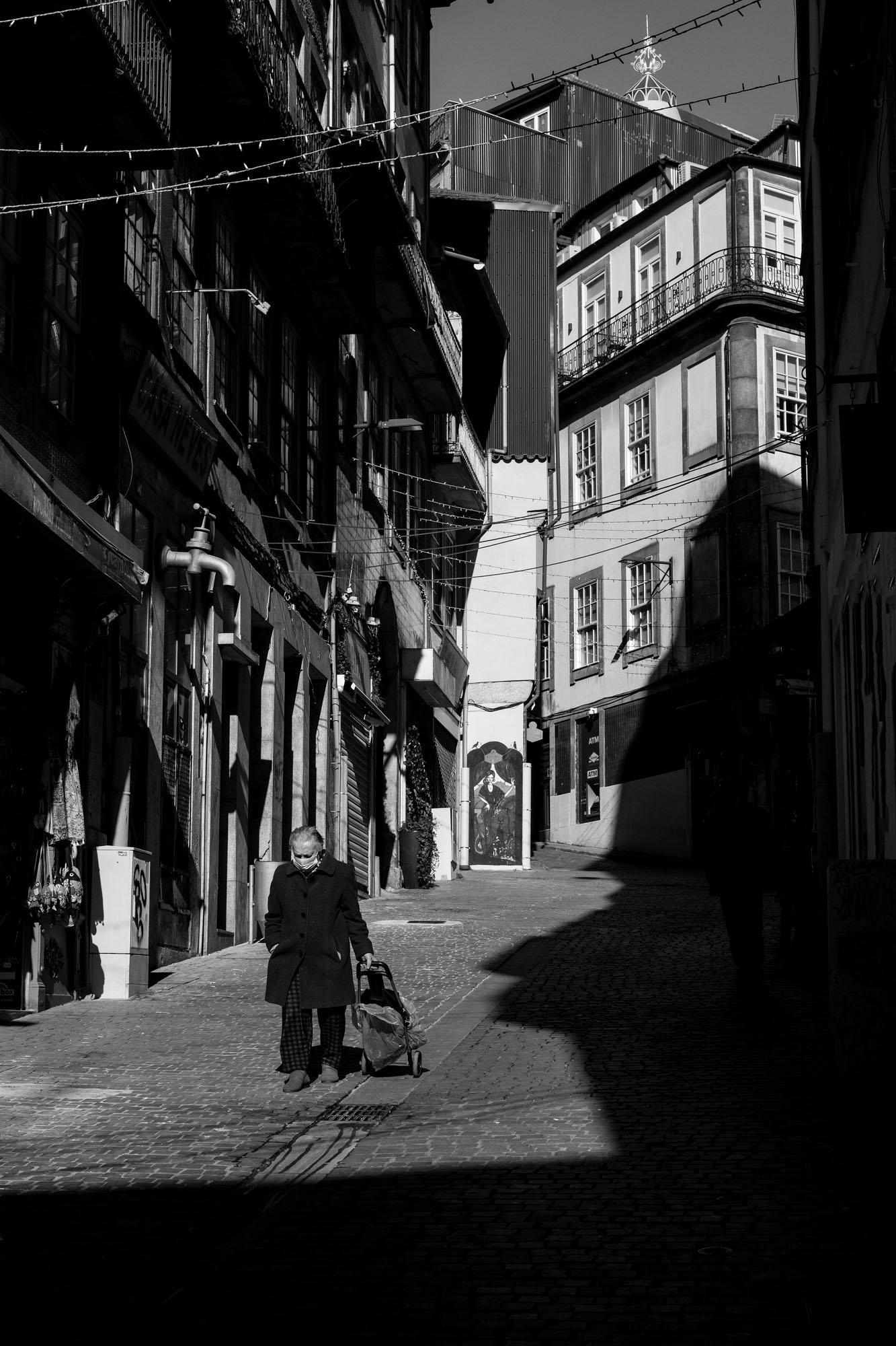  Older woman pulling a shopping trolley through a narrow street of light and shadow, Porto. Black-and-white street photograph by Chris Perona. 
