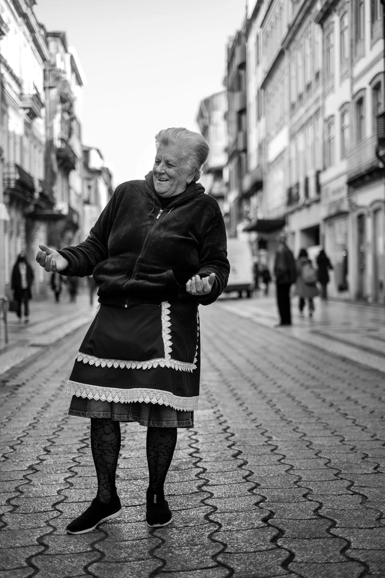  Smiling older woman in a traditional skirt dancing on a cobbled street, Porto. Black-and-white street portrait by Chris Perona. 