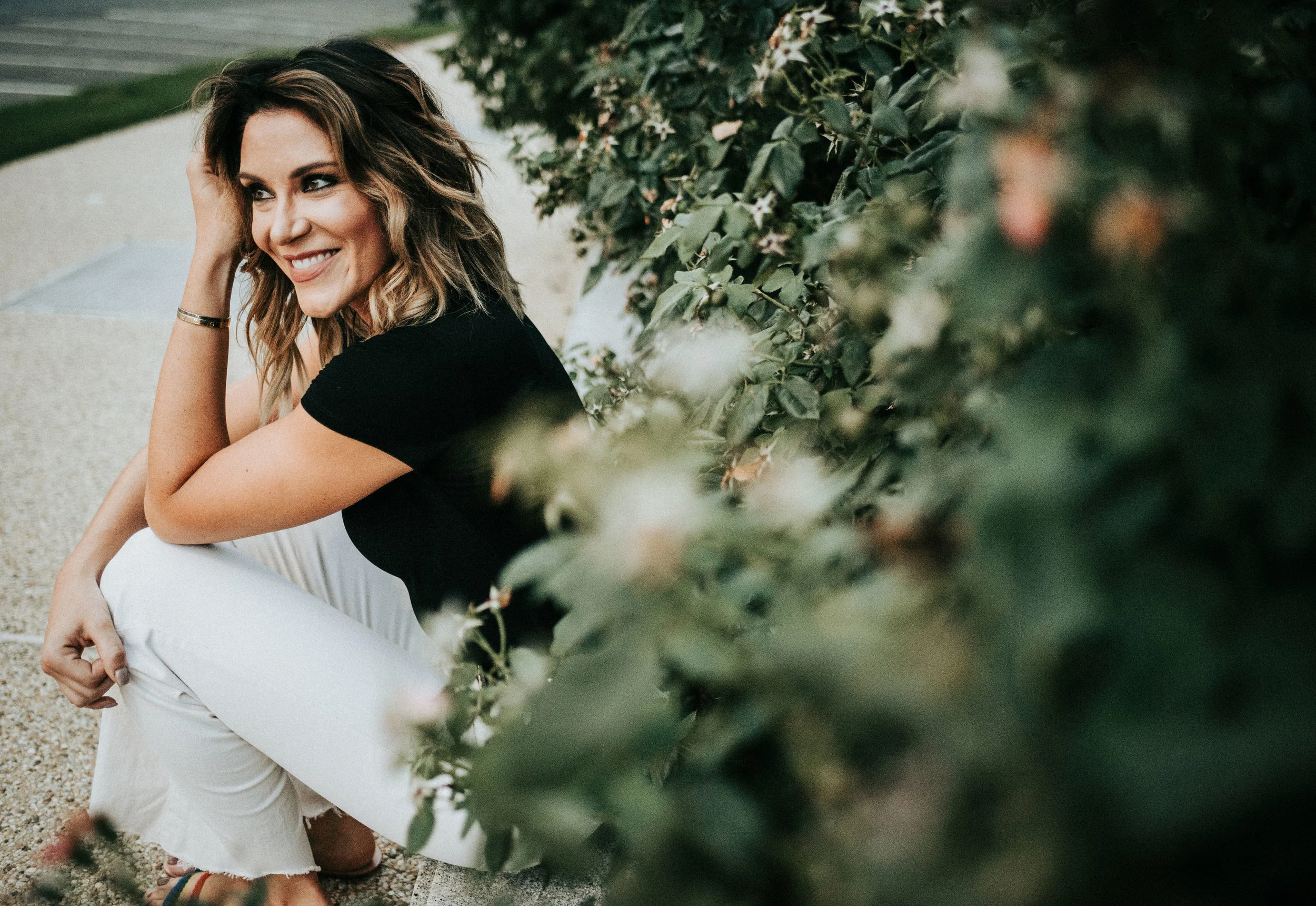 woman sits on sidewalk beside greenery smiling