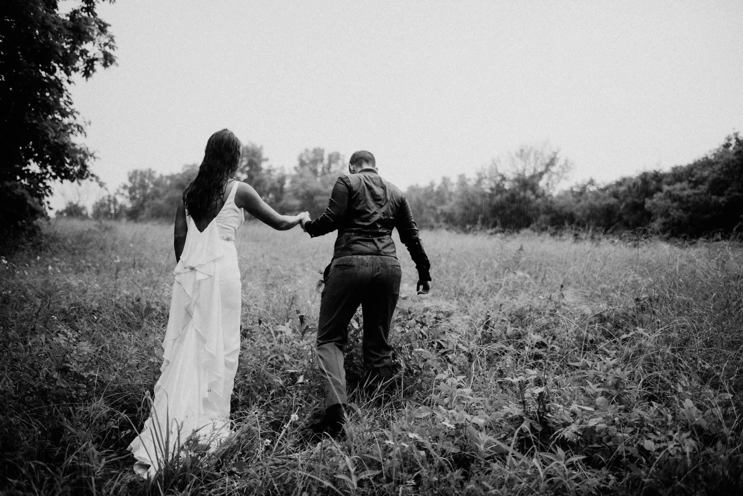 black and white photo of two married women in their wedding attire walking through field in the rain