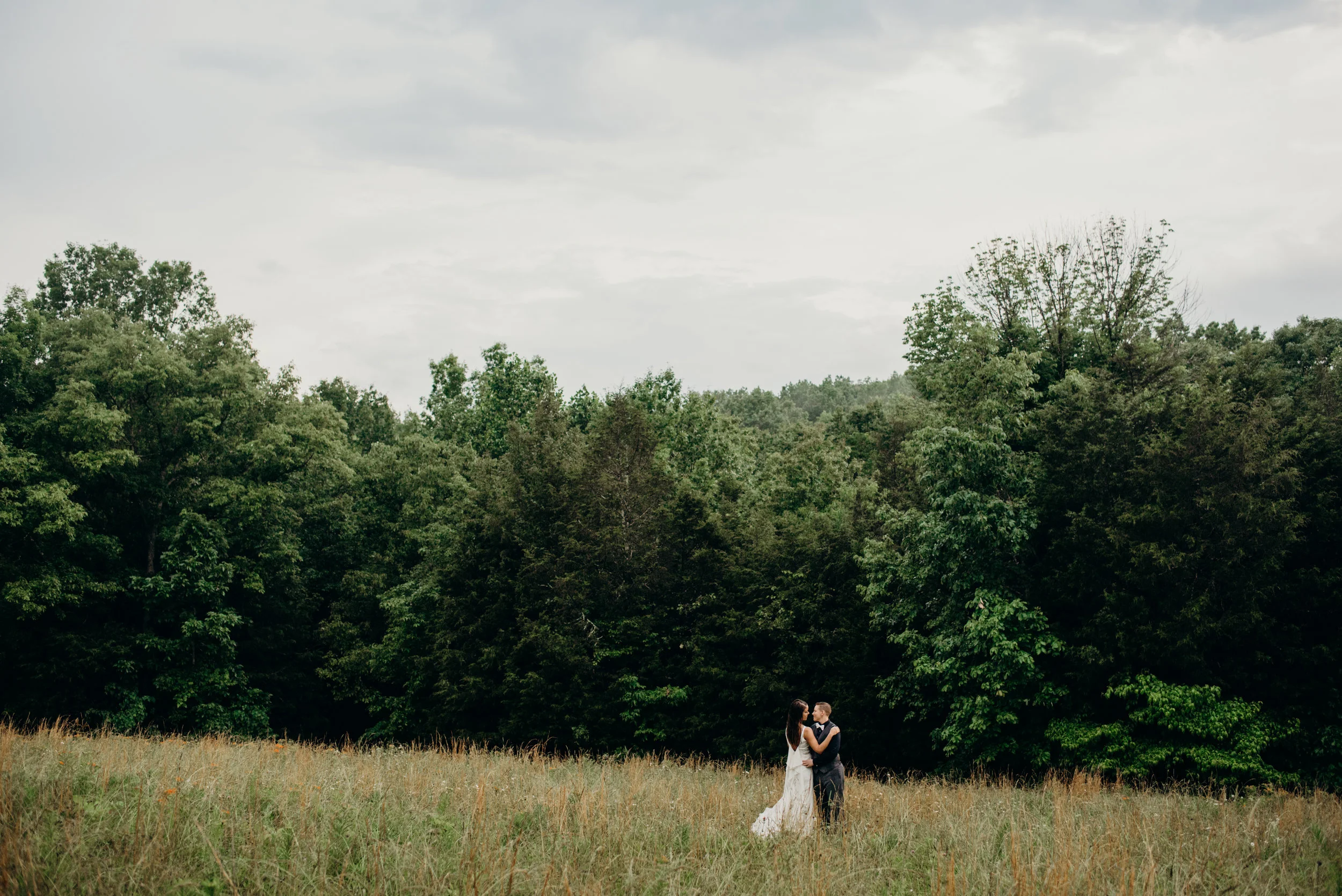 married couple in the distance embracing and looking at each other in a field surrounded by trees