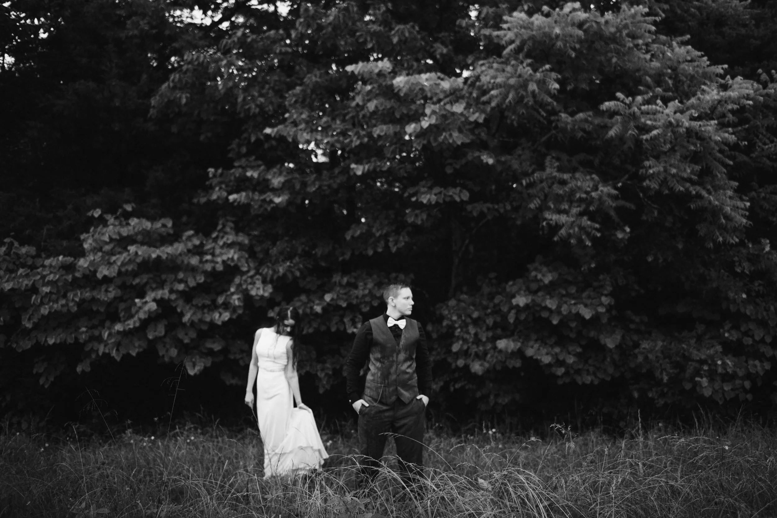 post-wedding session with two women posing in wedding attire in field with trees behind them