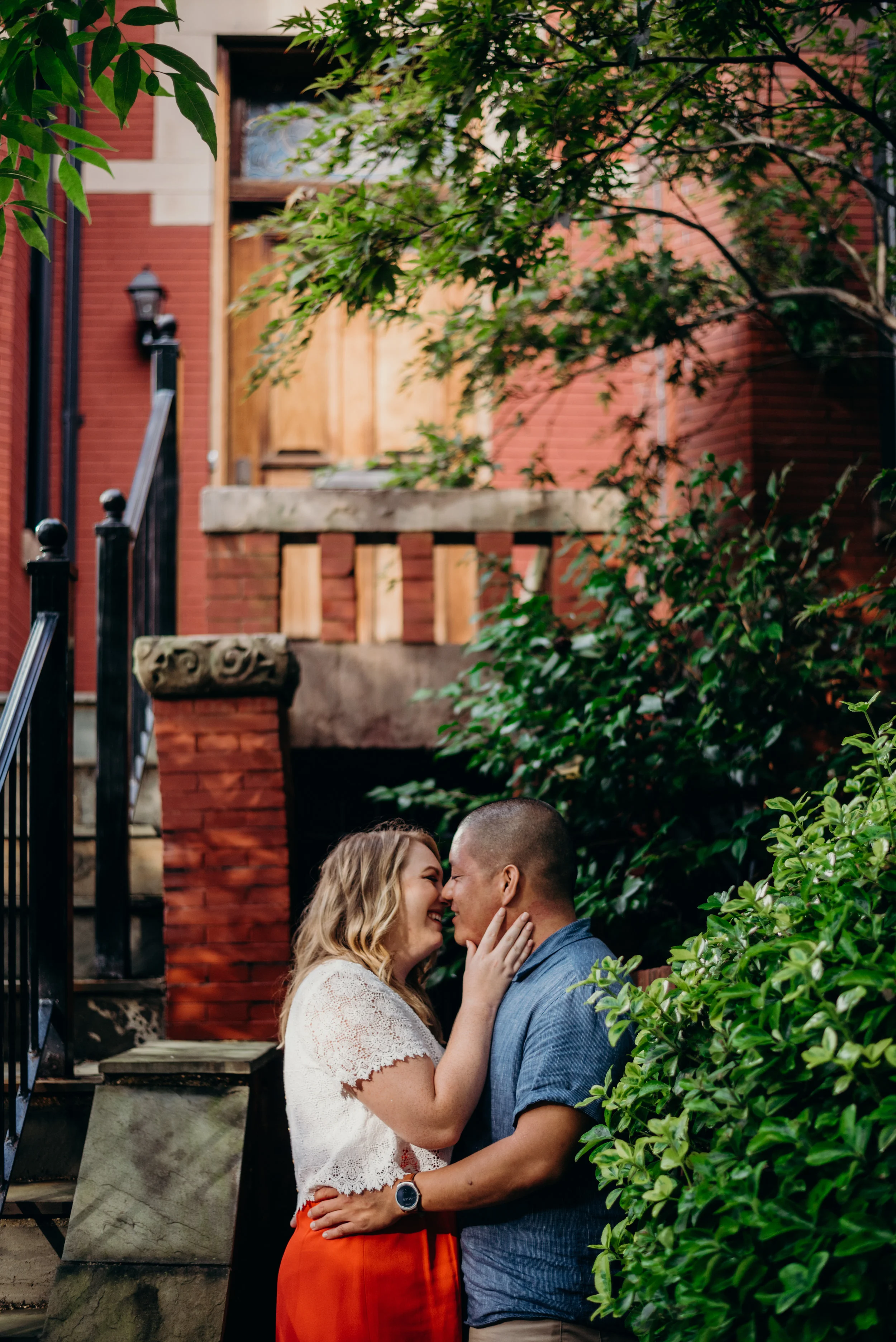 engaged couple kiss with brick building and trees in background