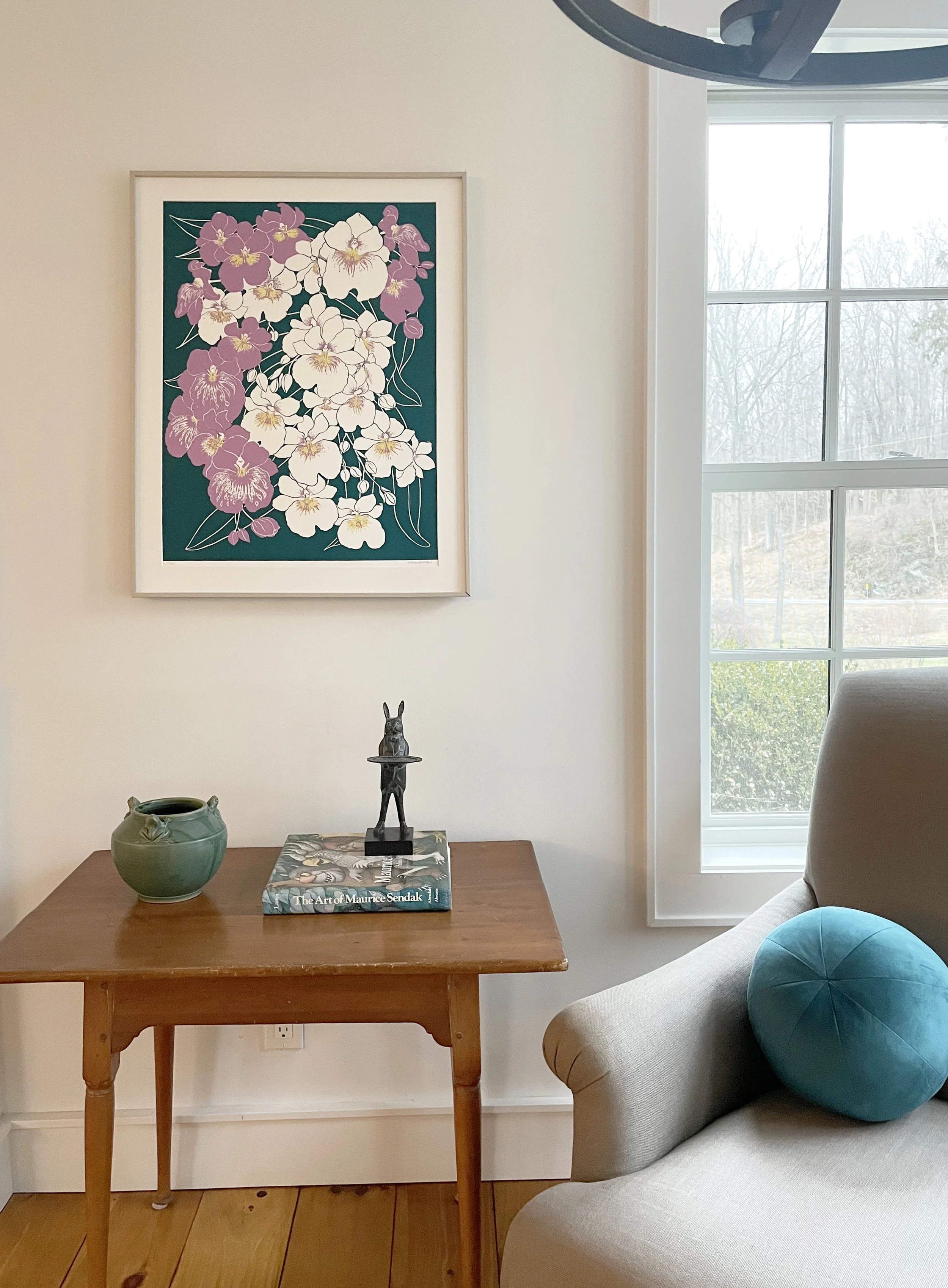 A cozy living room corner with a small wooden table, a green vase, a vibrant floral painting of white and purple orchids on the wall and a matching green velvet ball pillow on a cream armchair. 