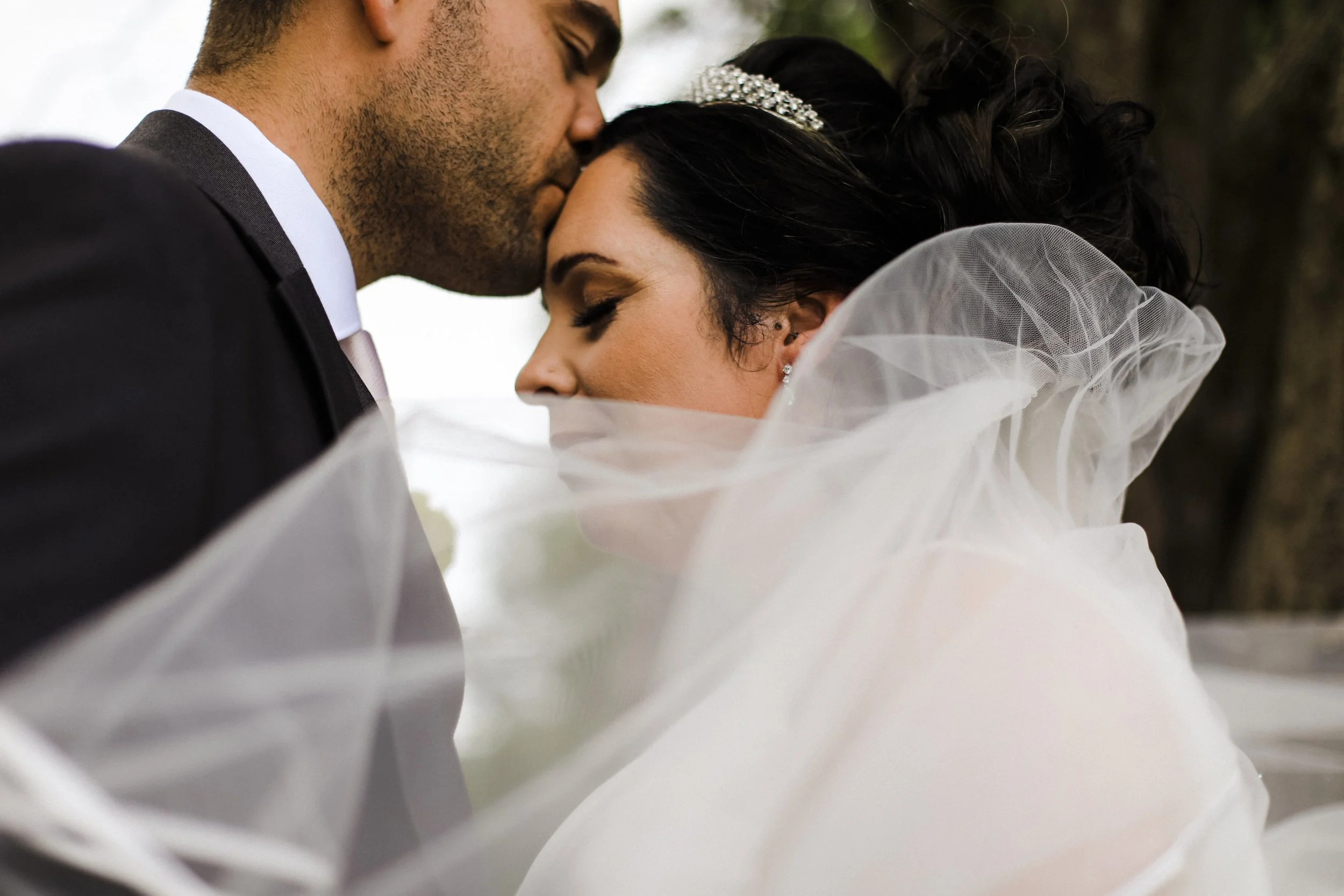 close up shot of groom kissing bride on the forehead with her veil blowing across them on the ground at Formby hall Liverpool Wedding Photographer.  