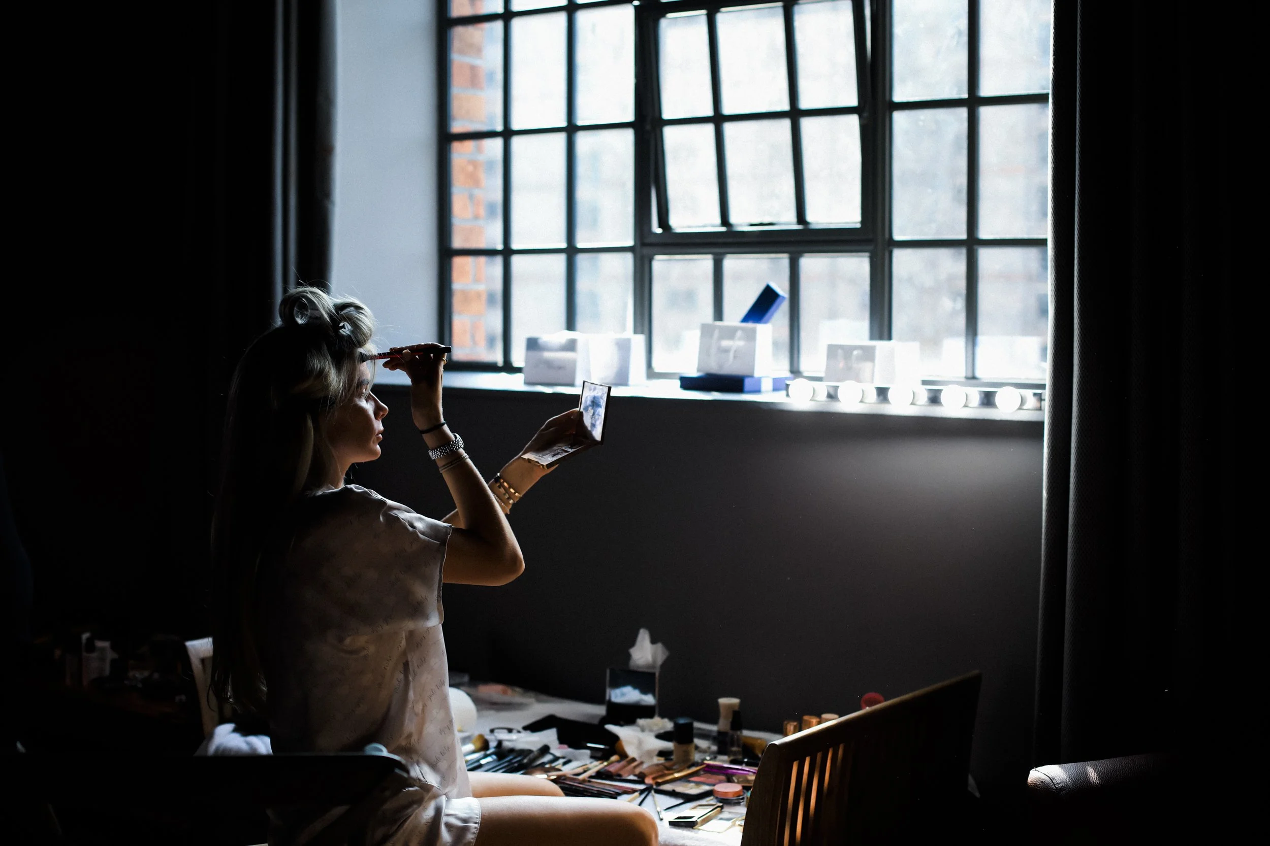 Independent Grace: Bridesmaid Sits Before the Window at Titanic Hotel, Skillfully Doing Her Own Makeup, Holding a Small Mirror, Adding a Touch of Self-Assured Elegance to the Titanic Hotel Liverpool W
