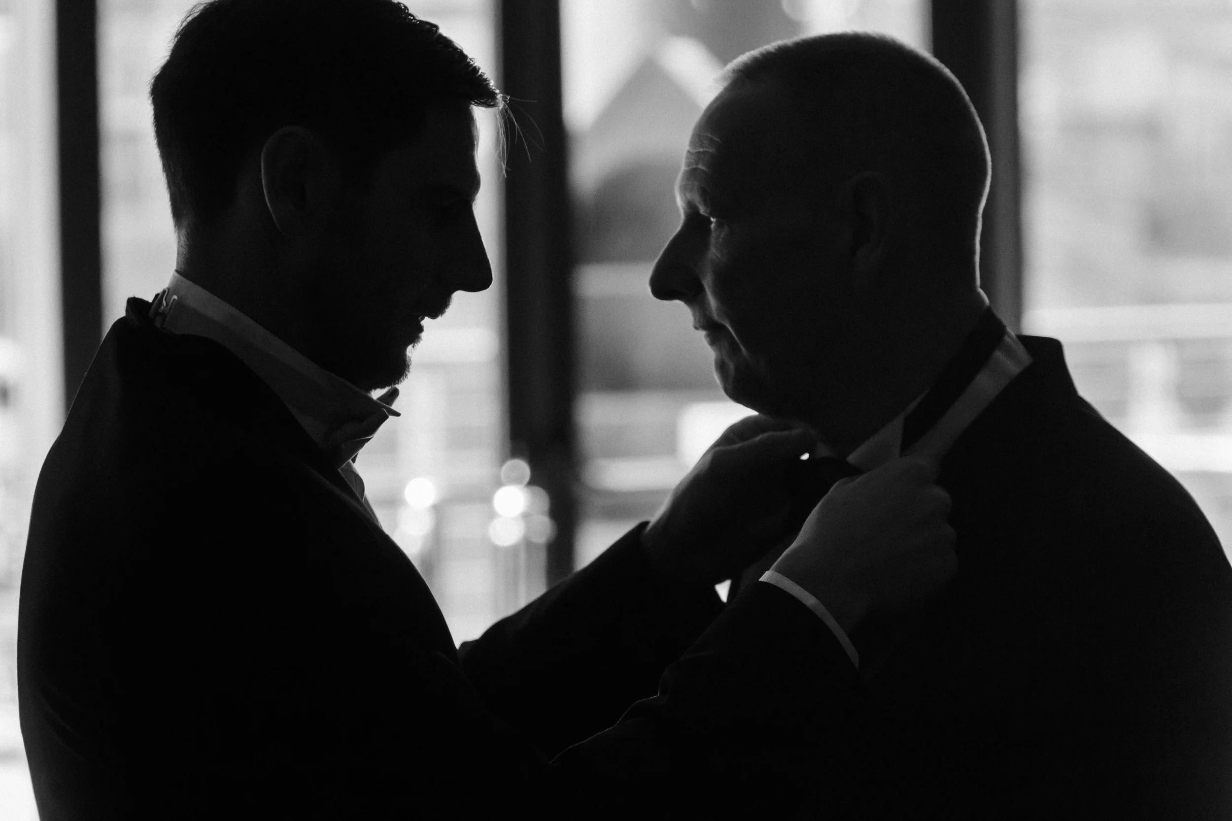 Generational Bond: Groom Assists in Tying His Father's Tie in the Titanic Hotel Lobby, Illustrating a Heart-warming Connection Between Father and Son on This Special Day.