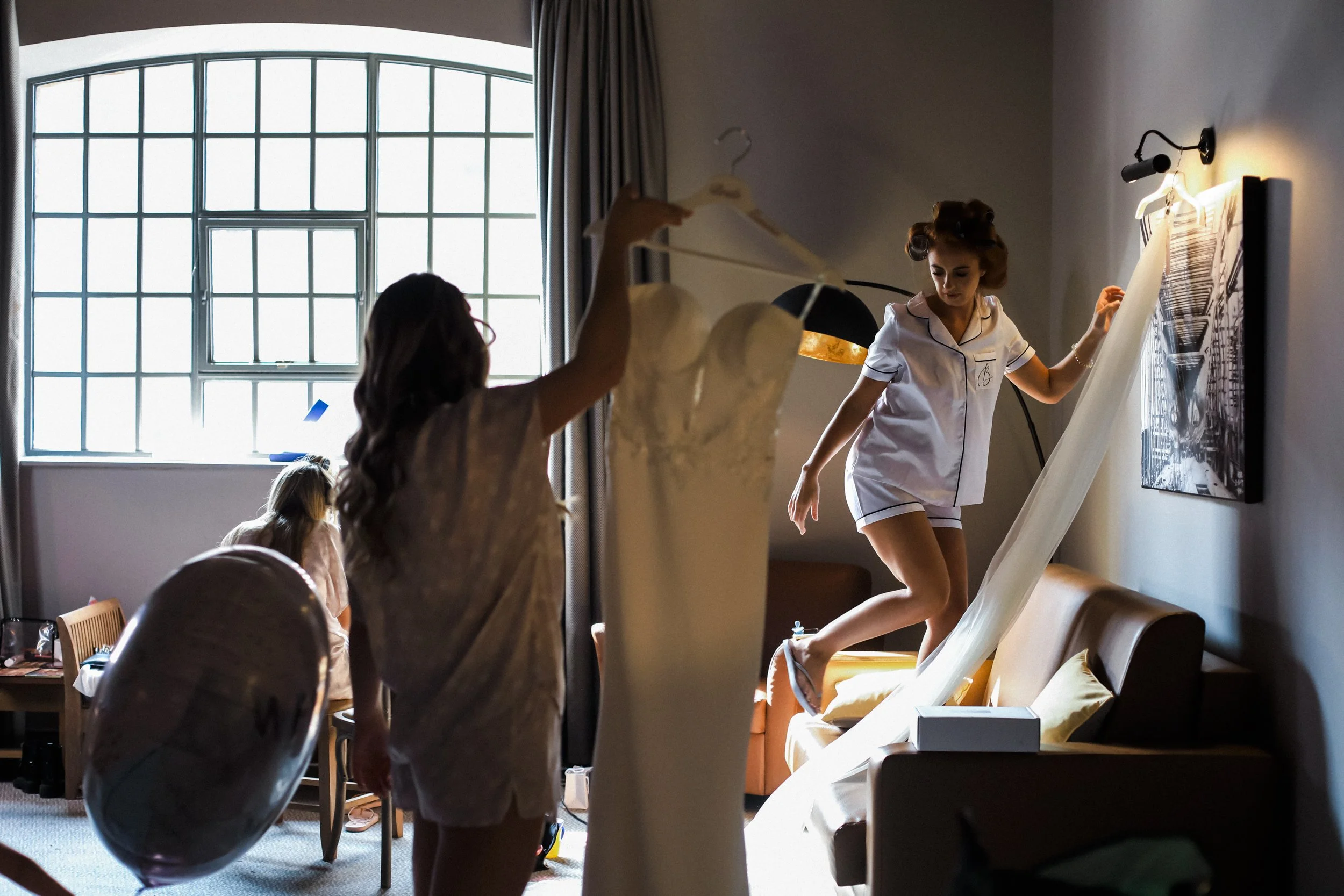  Graceful Preparation: Bride Climbs Down from the Couch After Hanging Her Veil, While Her Bridesmaid Holds Her Wedding Dress at Titanic Hotel Liverpool, Capturing a Moment of Poise and Collaboration.