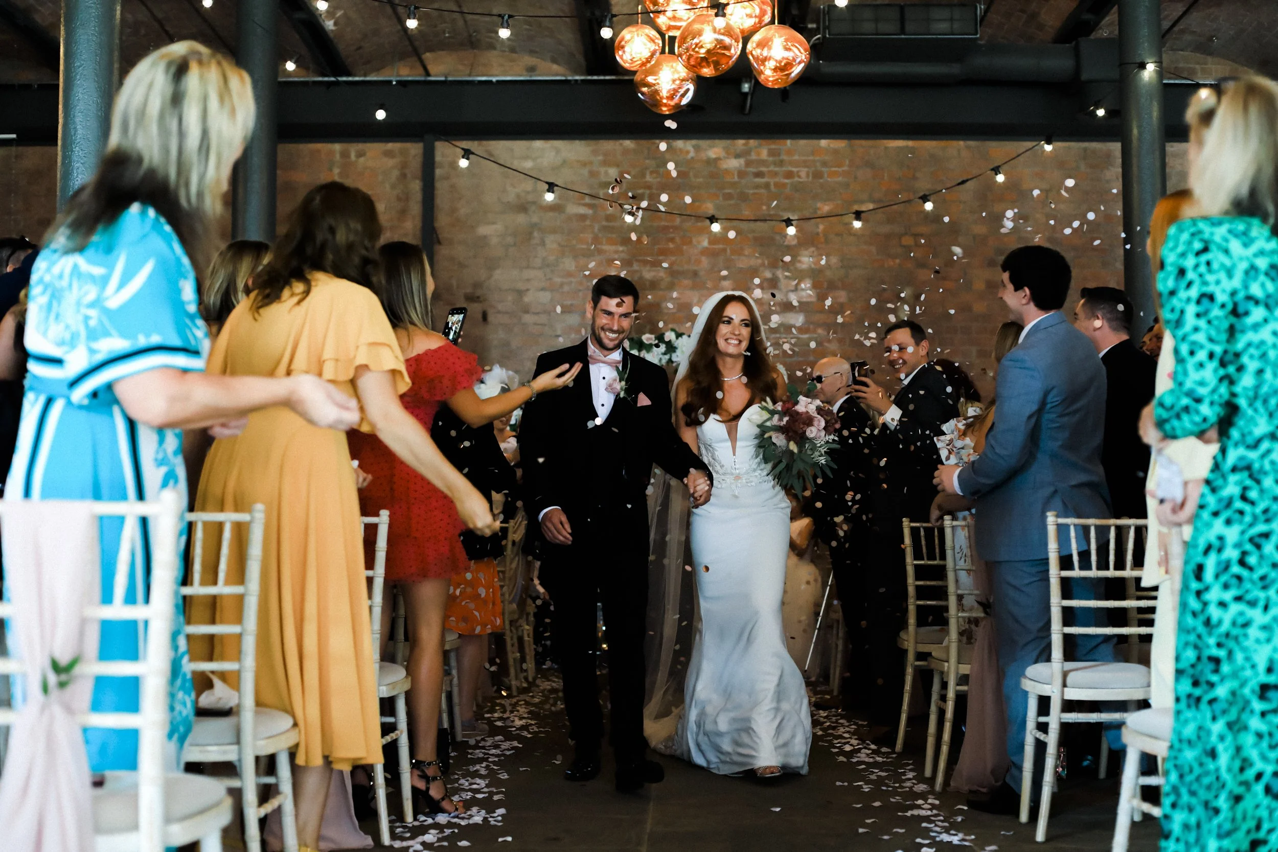 oyful Exit: Bride and Groom Leave the Ceremony Amidst a Shower of Confetti Tossed by Delighted Guests, Celebrating Their Union in a Blissful Moment.