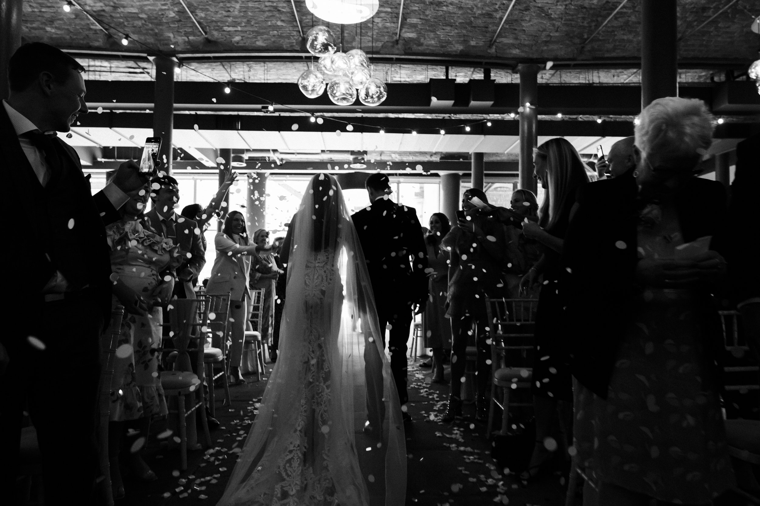 Picture-Perfect Exit: Wide Shot Captures Bride and Groom Leaving the Ceremony as Guests Create a Confetti Shower, Creating a Stunning Scene of Celebration and Happiness.