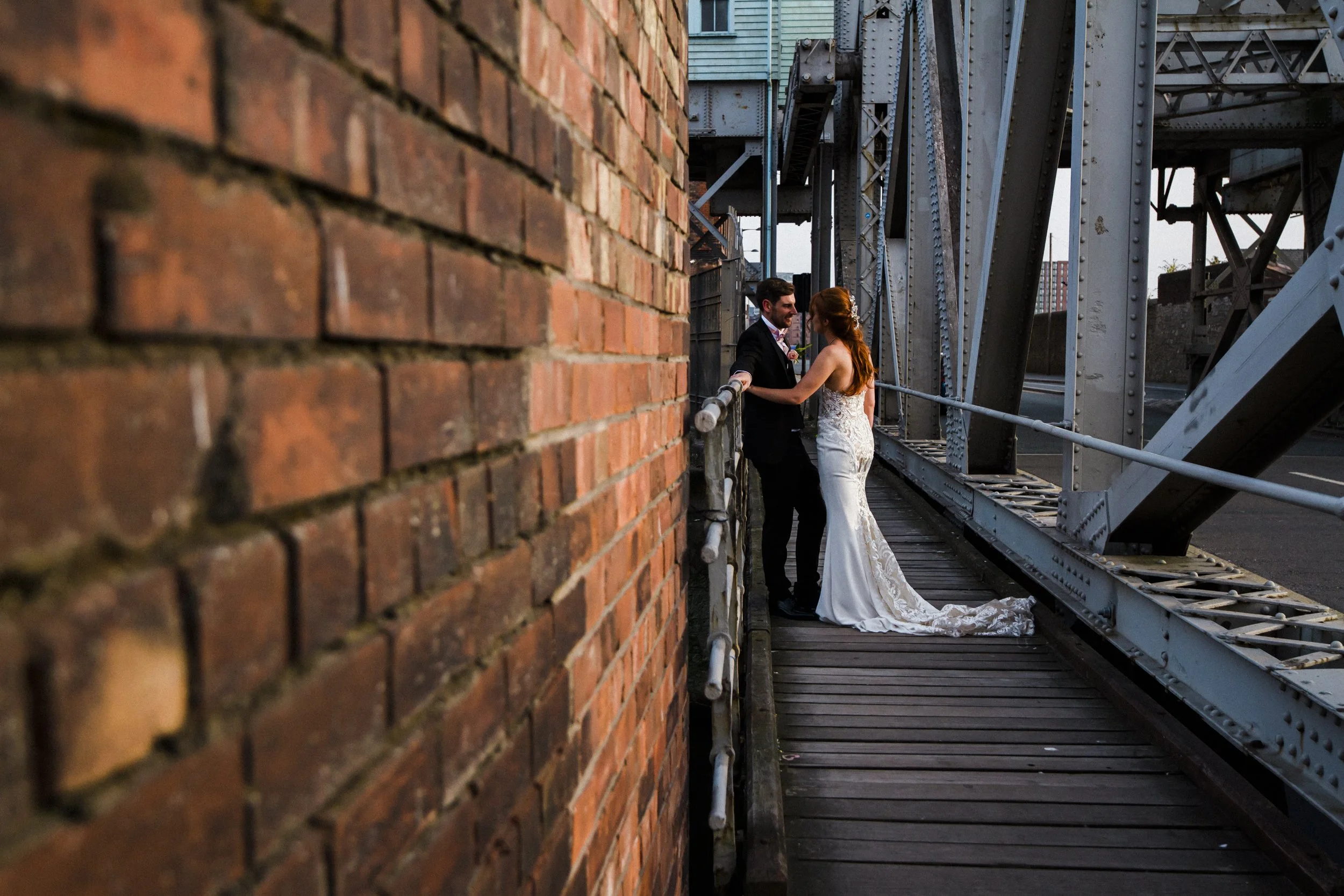 Intimate Harmony: Groom Leans on the Rail as Bride Stands Strong by Liverpool's Stanley Dock, Bathed in the Golden Hour Sunlight, Creating a Moment of Intimate Togetherness.