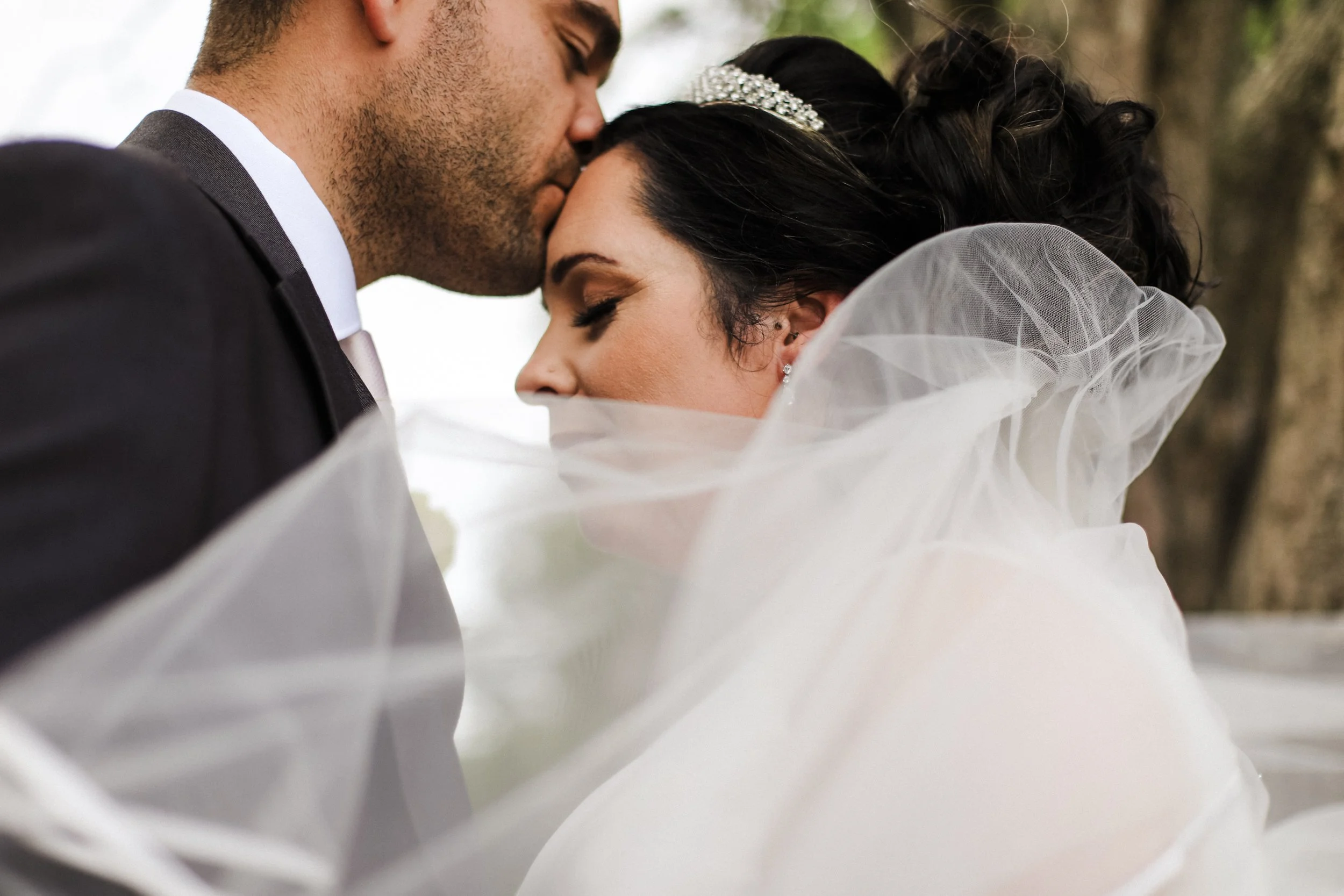close up shot of the groom kissing the bride on the forehead with her veil blowing across them on the grounds at Formby Hall Sefton Wedding Photography