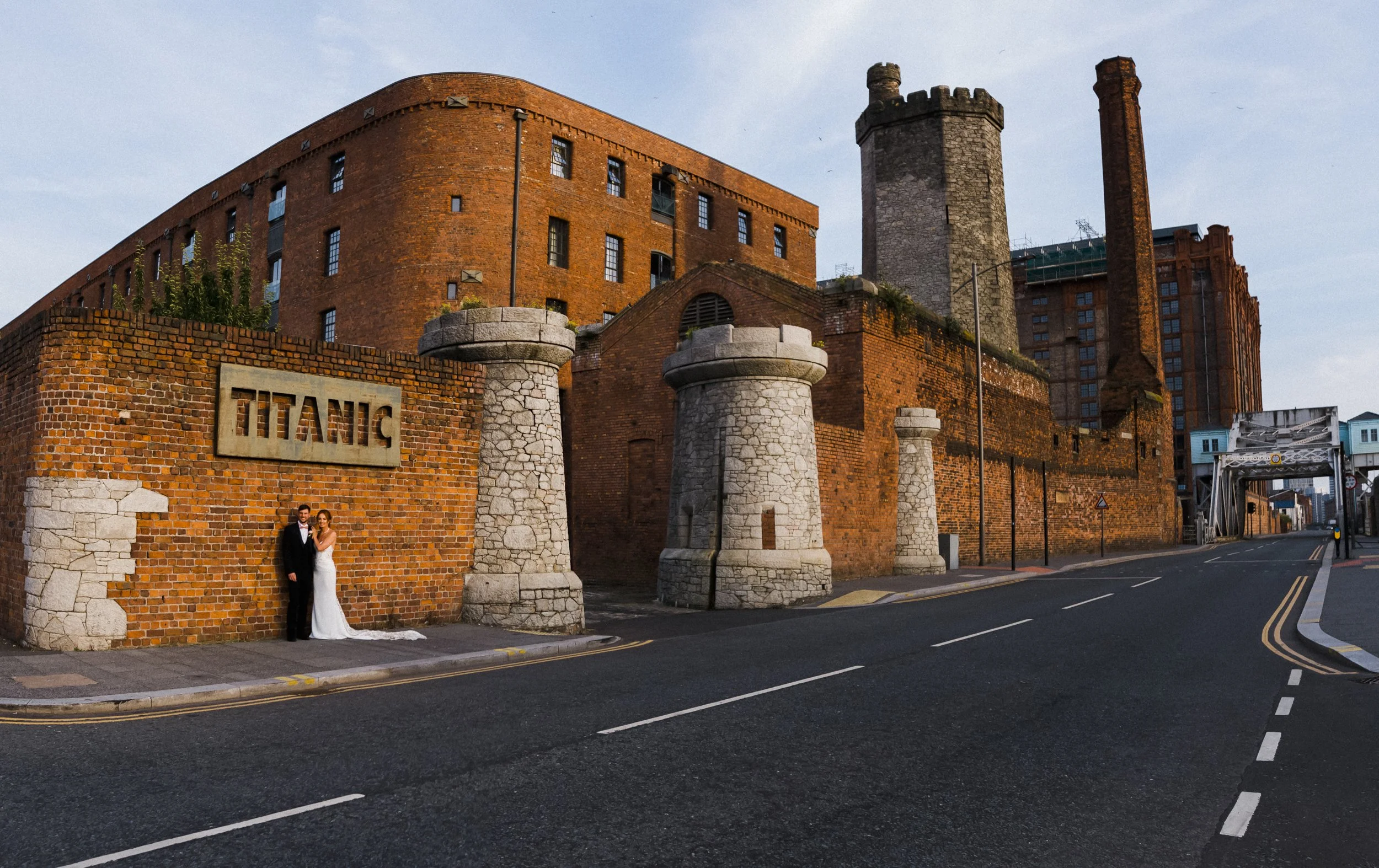wide shot of bride and groom standing under the Titanic hotel sign with the building and Stanley dock in golden hour sun light  Liverpool Wedding Photographer.