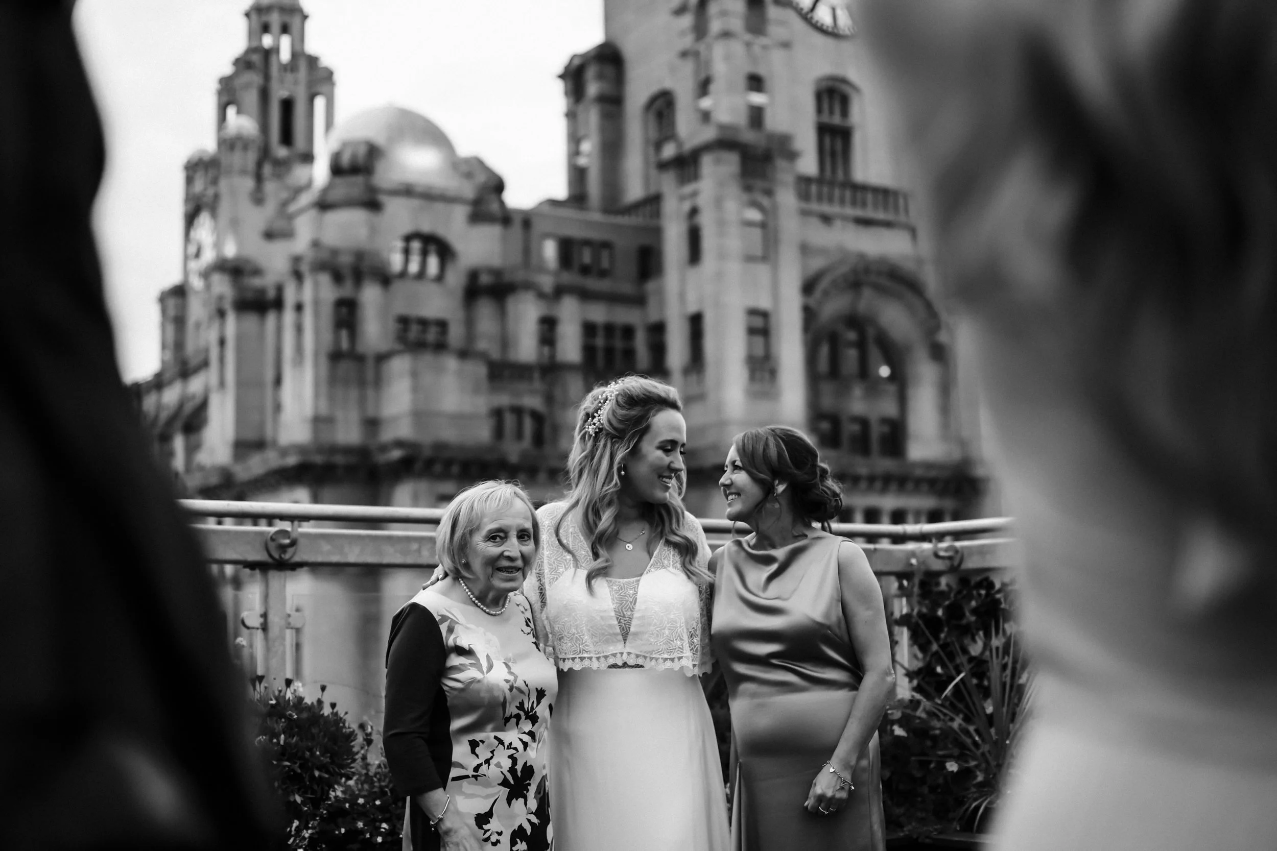 Candid moment on OH ME OH MY's rooftop: Bride, mum, and Nan share a heartfelt scene with Liverpool's iconic Liver Buildings in the background. Liverpool wedding photography perfection