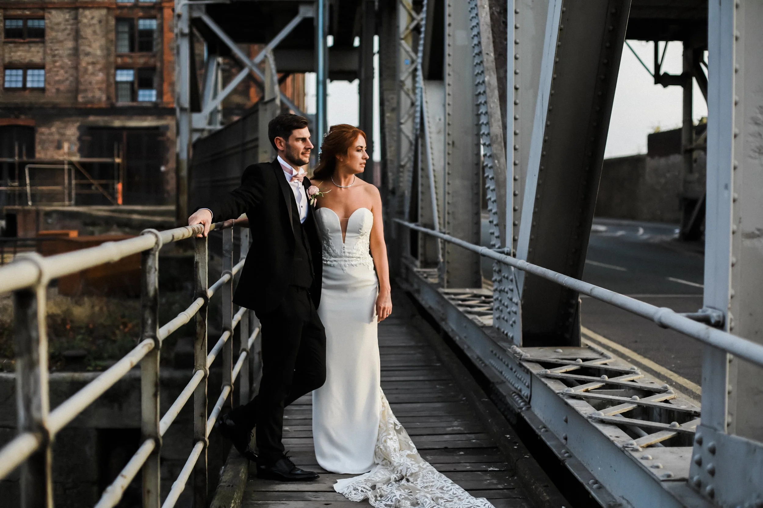 groom leans on rail and bride stands strong at Liverpool's Stanley Dock by the Titanic Hotel in golden hour sunlight Liverpool Wedding Photography