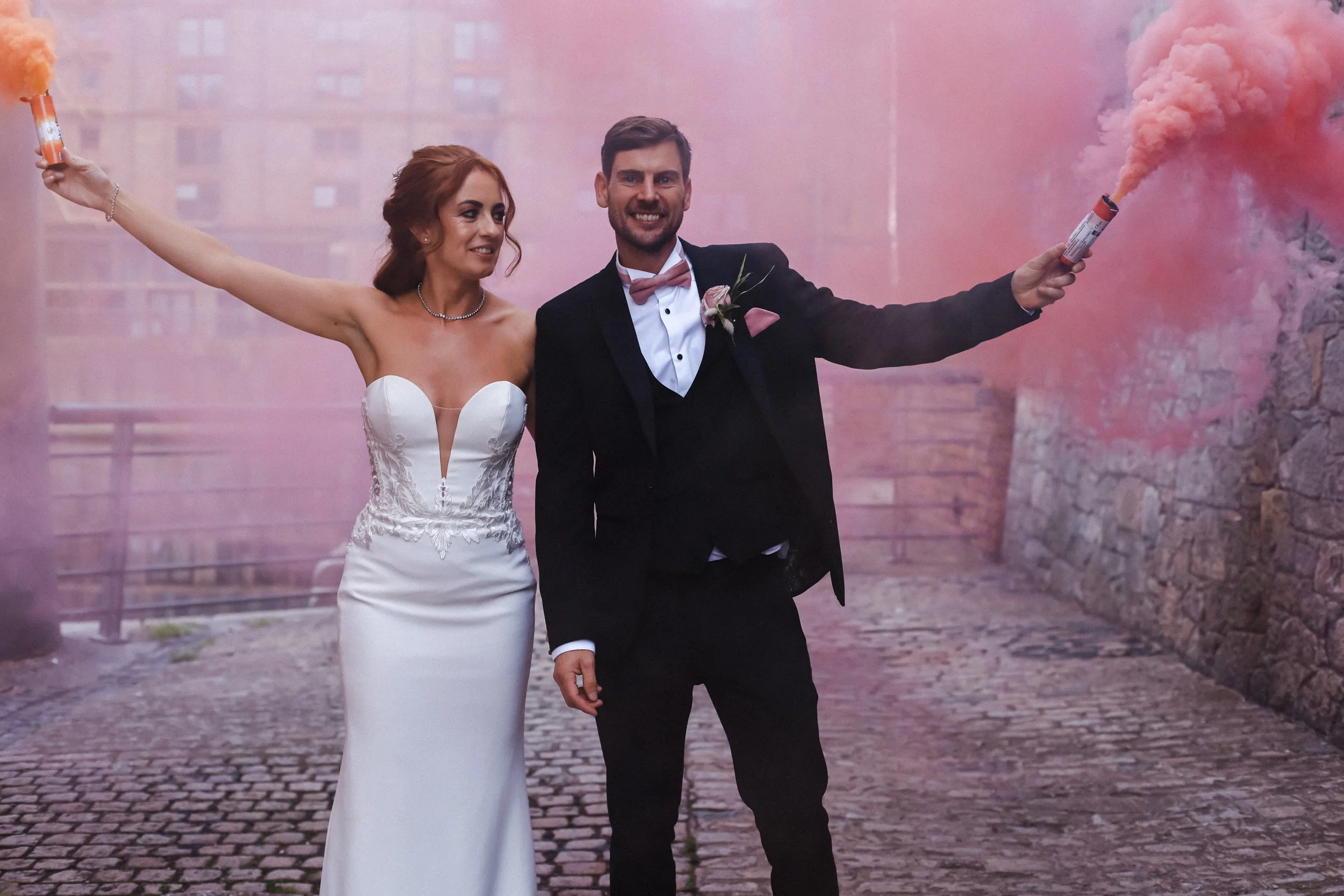 Bride and groom enjoy playful moments with pink and orange smoke bombs at Titanic Hotel, creating vibrant and lively scenes in this Liverpool wedding photography capture.