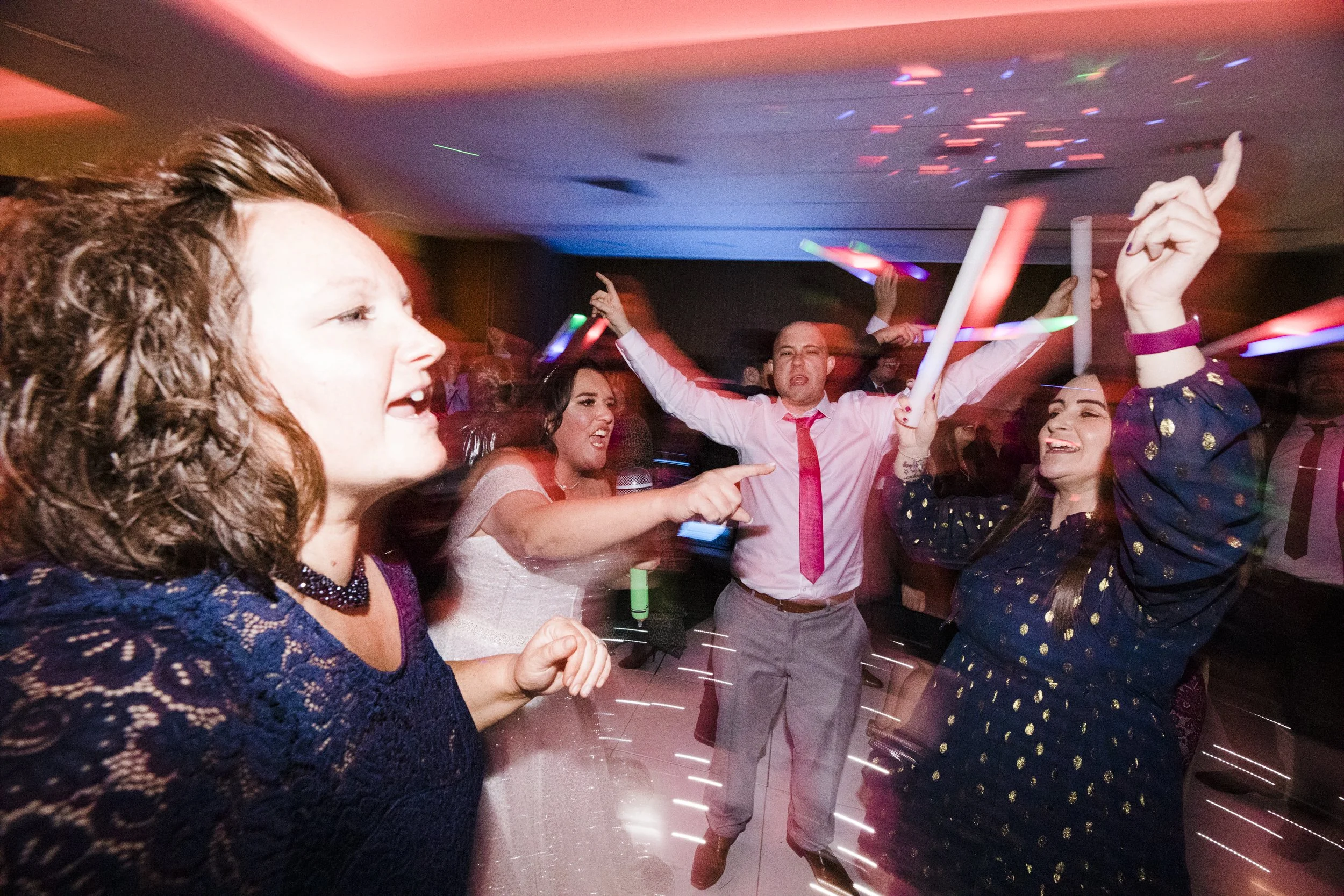 Bride and guest joyfully share a vibrant moment on the dance floor at the Holiday Inn Hotel, captured in a lively, full-rave mood. The dynamic shot pulsates with energy, radiating celebration