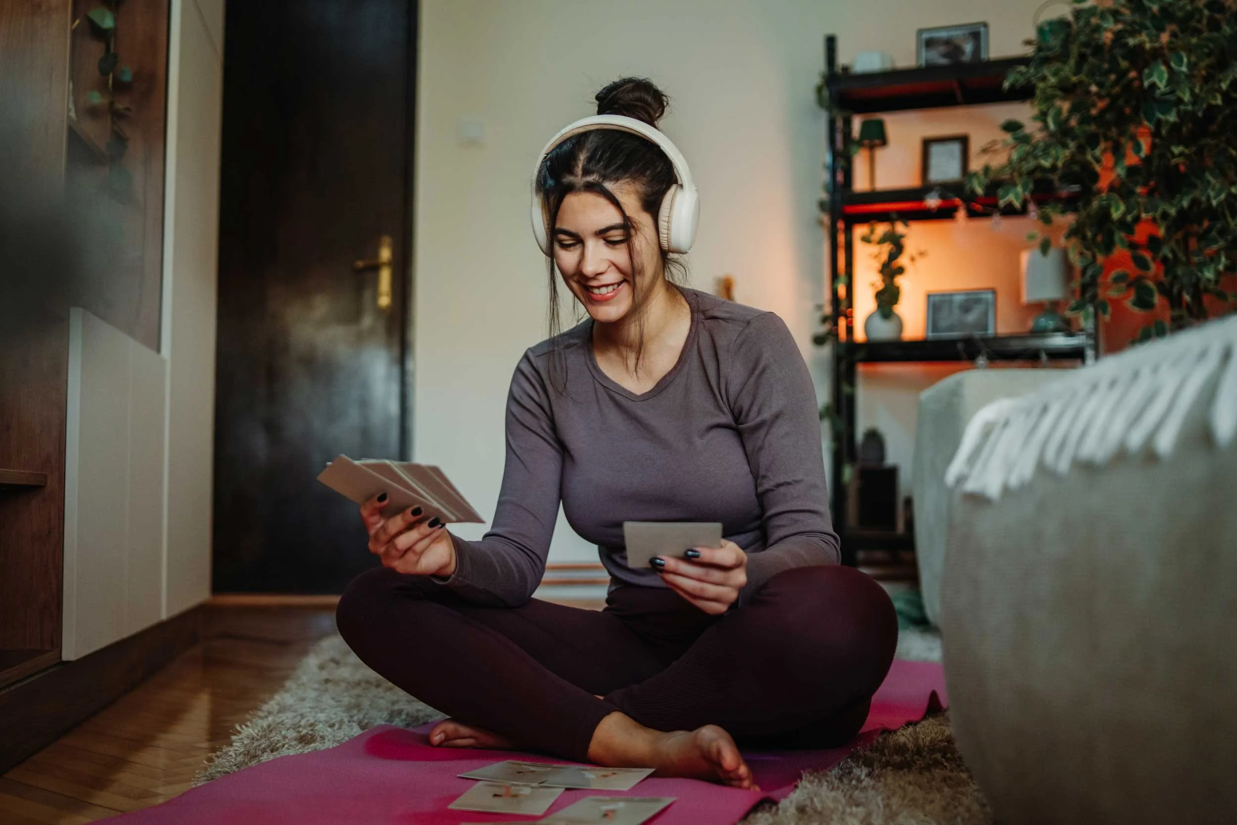 A young woman wearing headphones and sitting on a mat on the floor studies tarot.