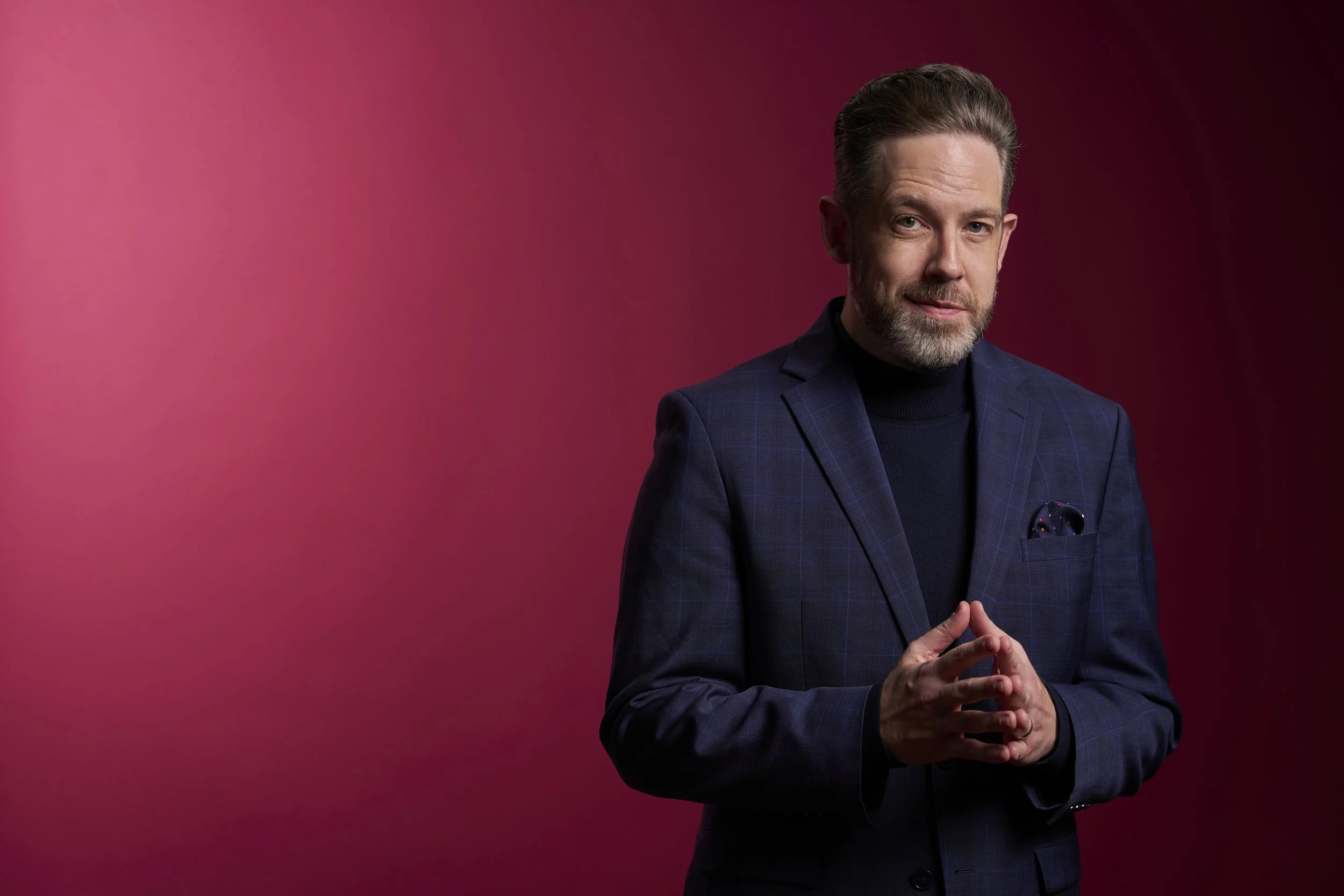 Brett Mitchell, American conductor, composer, and pianist, in a professional portrait dressed in a dark suit with a checkered pattern and a blue turtleneck, with his hands clasped in front of him.