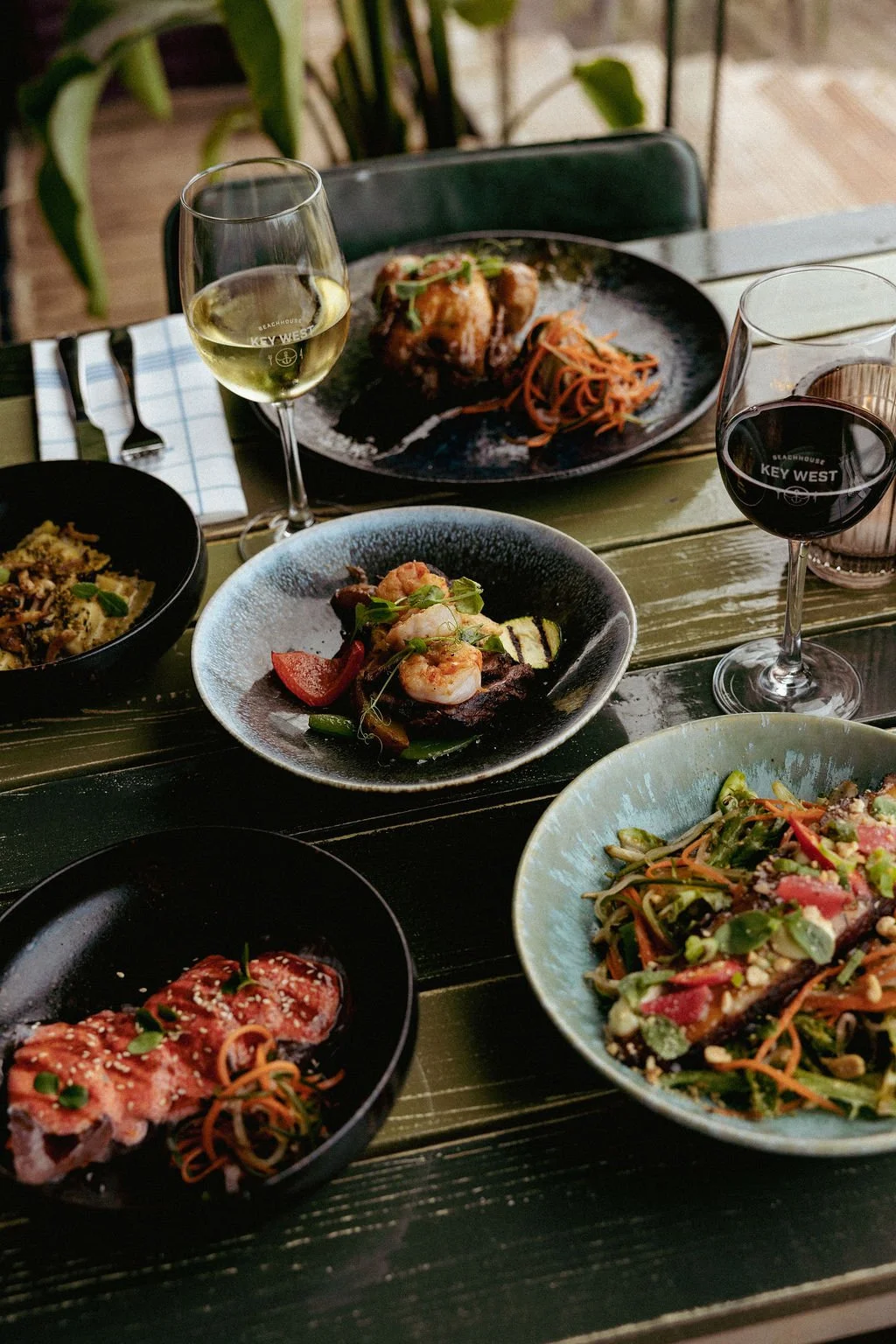 Een dinertafel met diverse gerechten, waaronder een bord met een salade met shrimps en groenten, een plaat met vlees en garnituren, een glas witte wijn, een glas rode wijn, en een ander gerecht met pasta en vlees op een houten tafel. In de achtergron