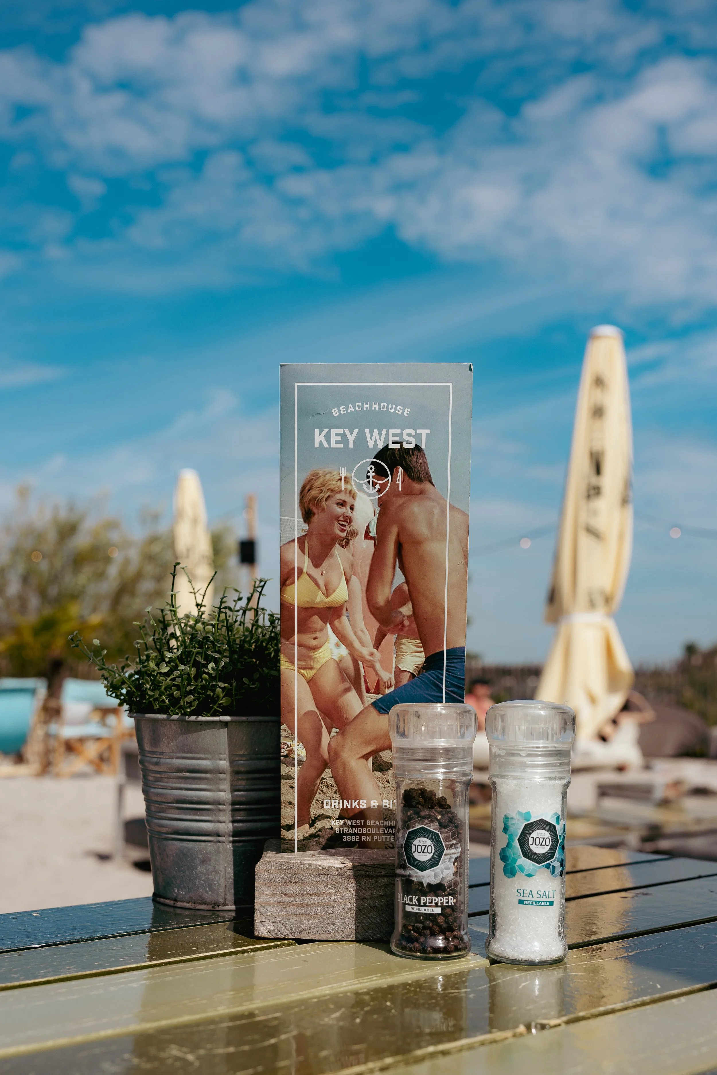 Sommertafel op strand met piment, zeezout en een menu met foto van twee vrouwen en een man in badkleding.