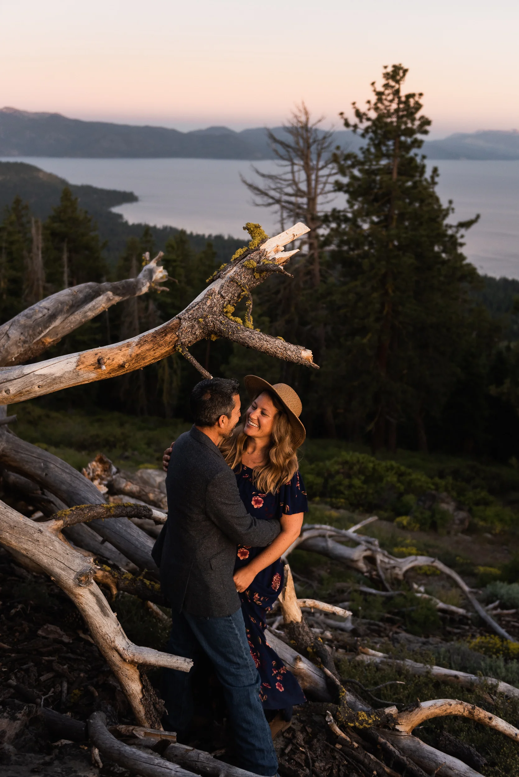 A couple looks each other in the eyes, smiling big, holding each other close. They’re high on a hillside overlooking Lake Tahoe during an anniversary session. They’re tucked up near a fallen tree, whose branches like tendrils crawl this way and that. Trees, mountain, and Tahoe are in the background.