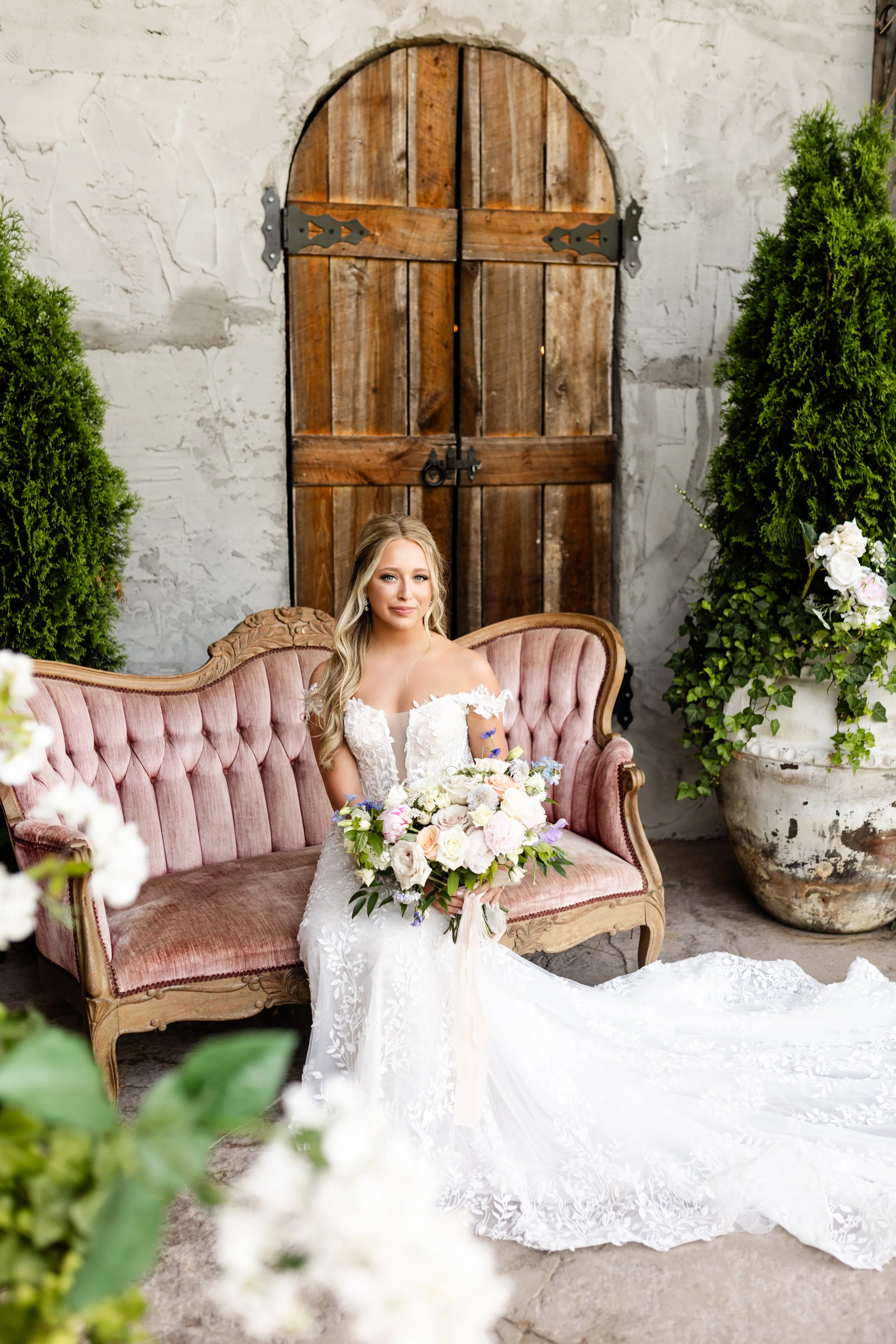 A bride sitting on a vintage pink cushioned sofa holding a large bouquet of flowers in front of a rustic wooden door and greenery.