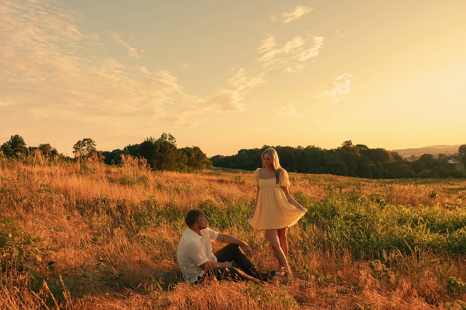 A man sitting on grass in a field with a woman standing near him, both enjoying sunset in a rural landscape.