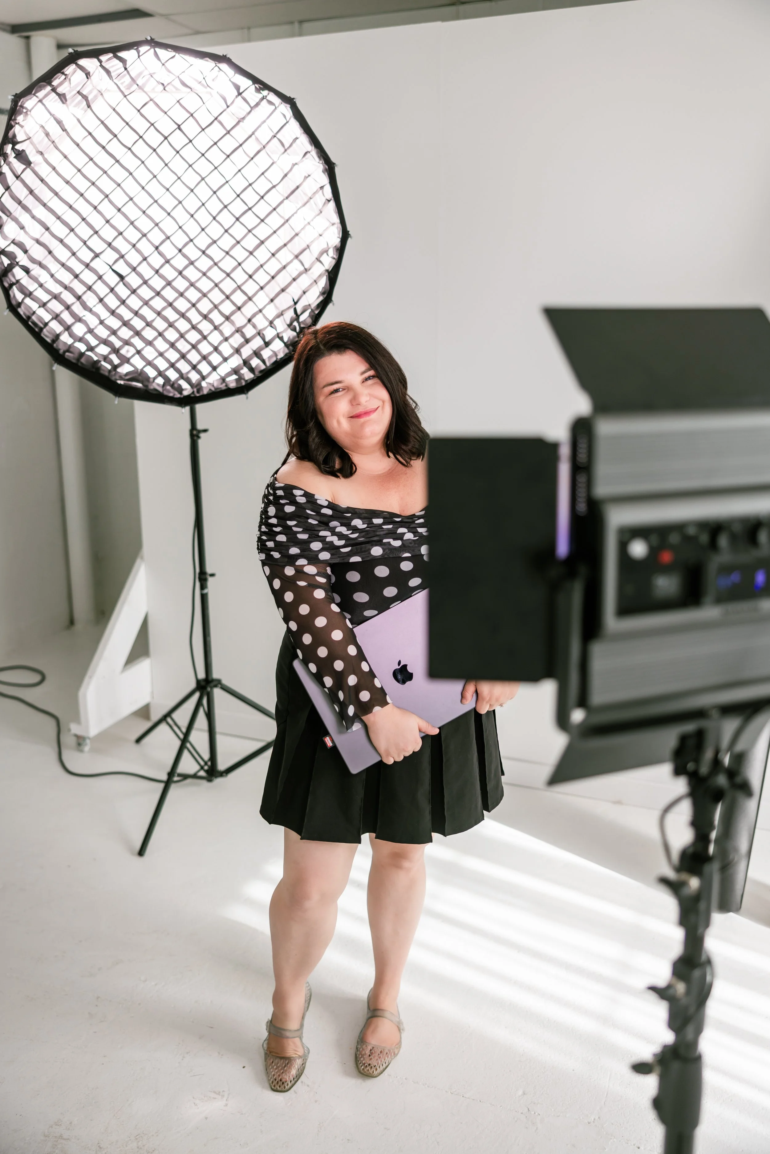 A woman standing in a photography studio, holding a silver laptop, smiling, near studio lighting equipment, wearing a black and white polka dot top with sheer sleeves, black skirt, and heels.
