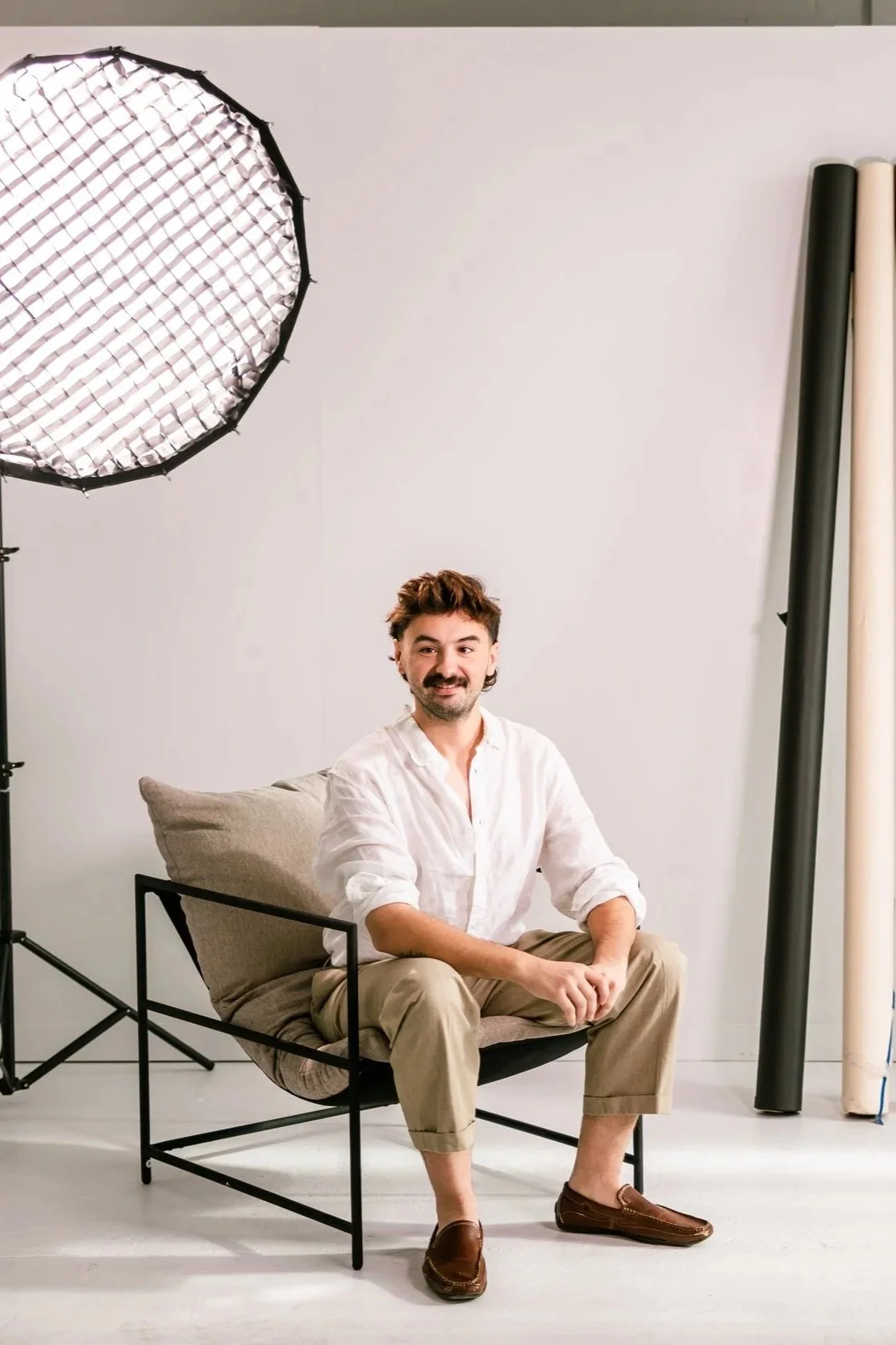 A man with brown hair and a beard, wearing a white shirt and beige pants, sits on a modern black chair with a beige cushion, smiling at the camera in a photography studio with a large professional light on the left and rolls of backdrop paper on the right.