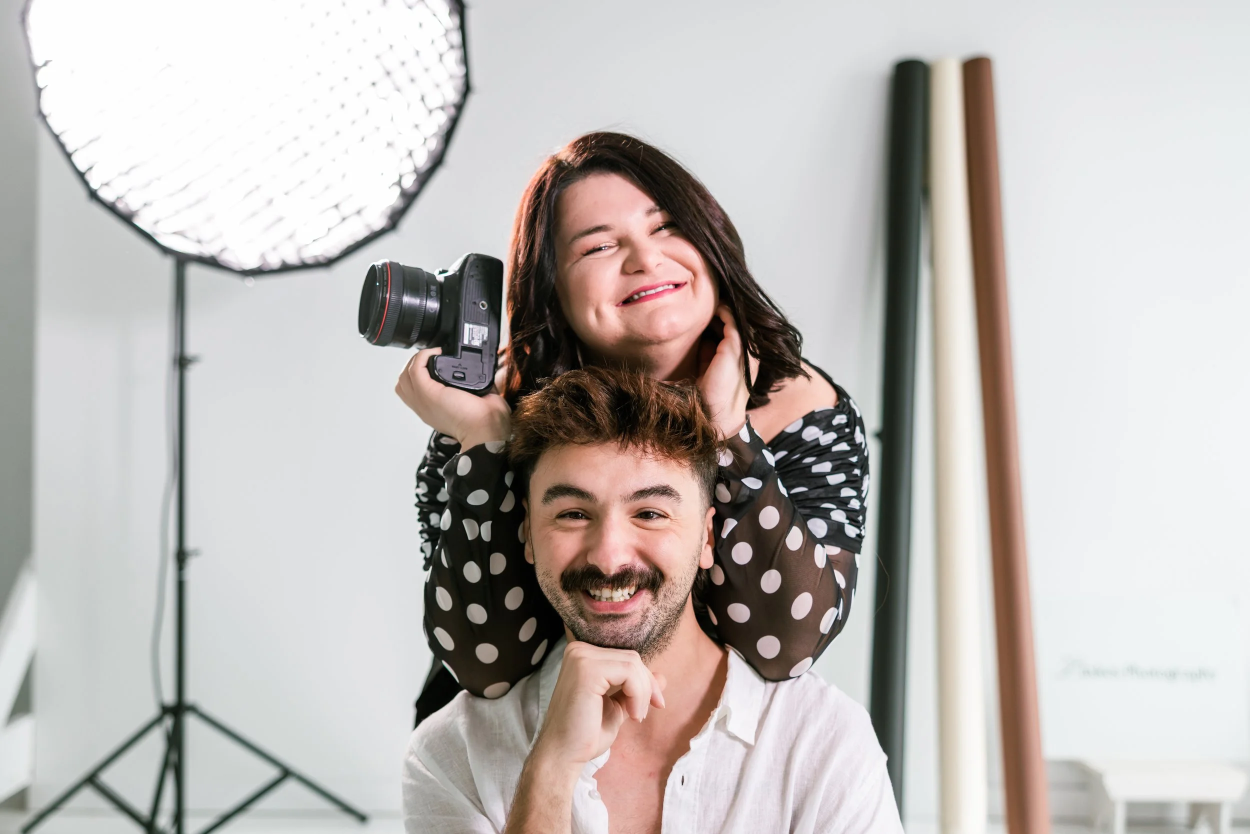 A woman with dark hair and a woman with brown hair and a beard, both smiling, with the woman holding a camera, in a photography studio with professional lighting equipment.