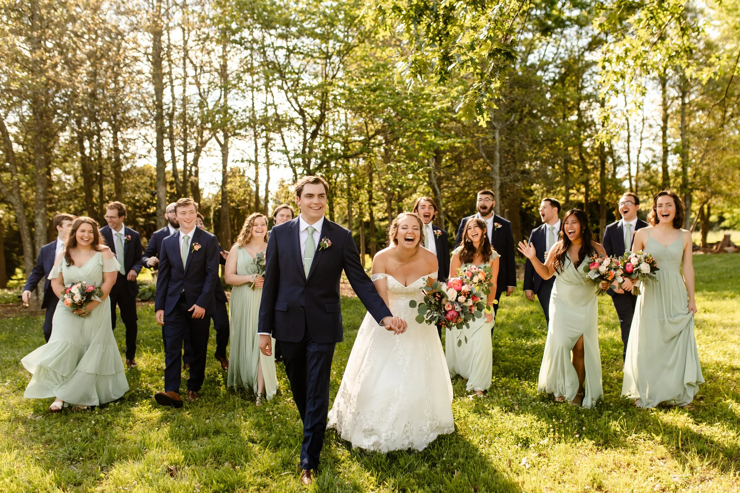 A wedding party walking outdoors on a sunny day, with the bride and groom in the center. The bride is in a white wedding dress holding a bouquet, and the groom is in a navy suit with a light green tie. Bridesmaids in pastel green dresses and groomsmen in navy suits are smiling and laughing, surrounded by green trees and grass.