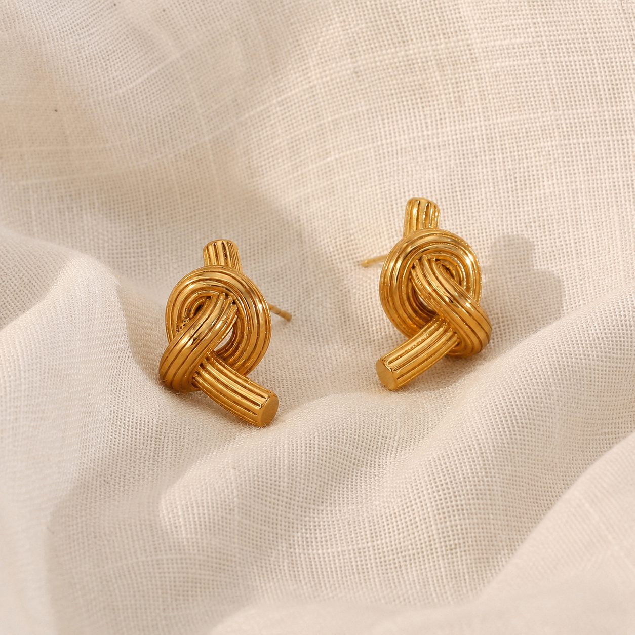 Close-up of gold knot stud earrings with ribbed rope detailing and a high-shine finish, displayed on natural cream linen.