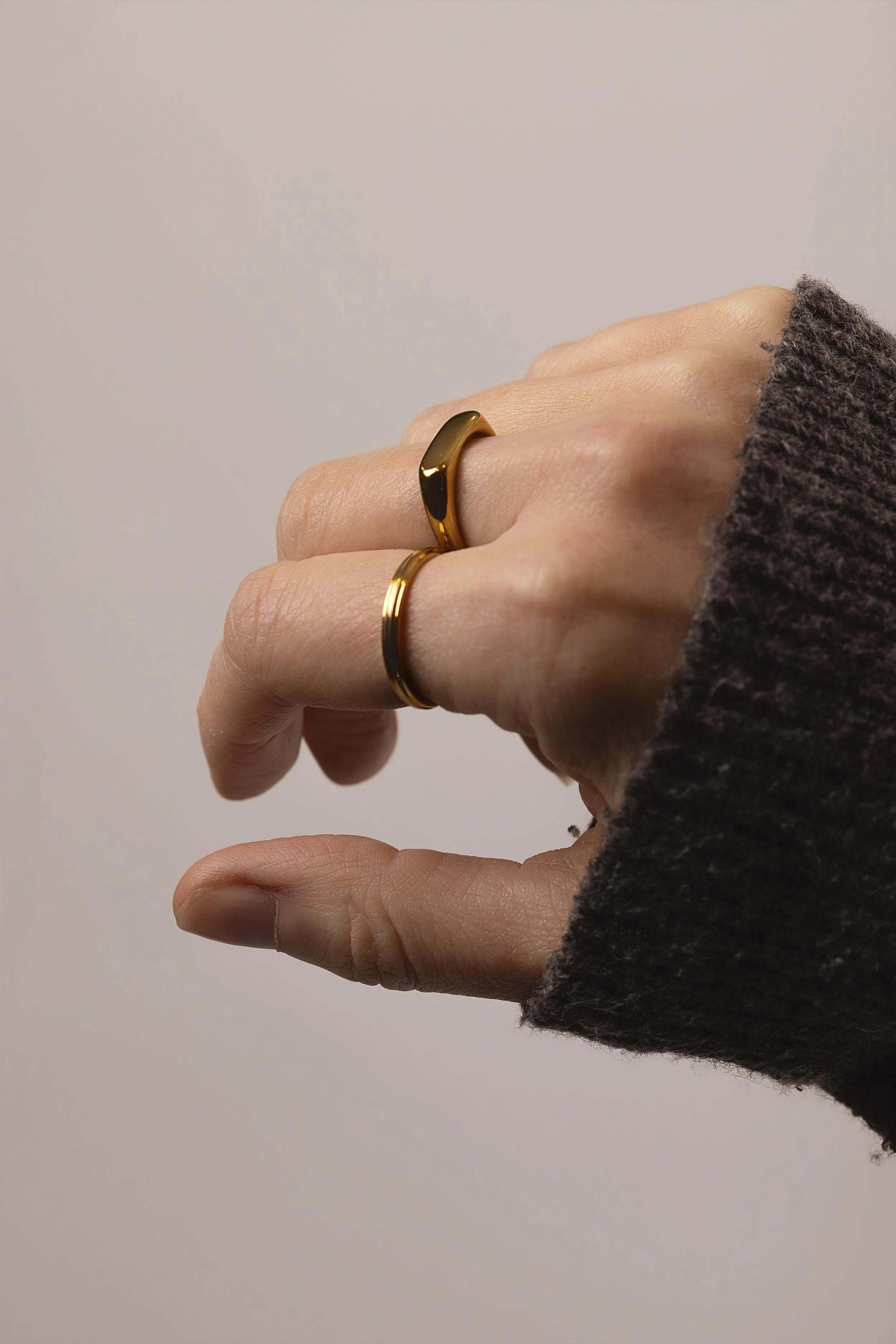 Model wearing slim gold signet ring stacked with a thin gold band ridge ring on index finger, styled with a grey knit jumper