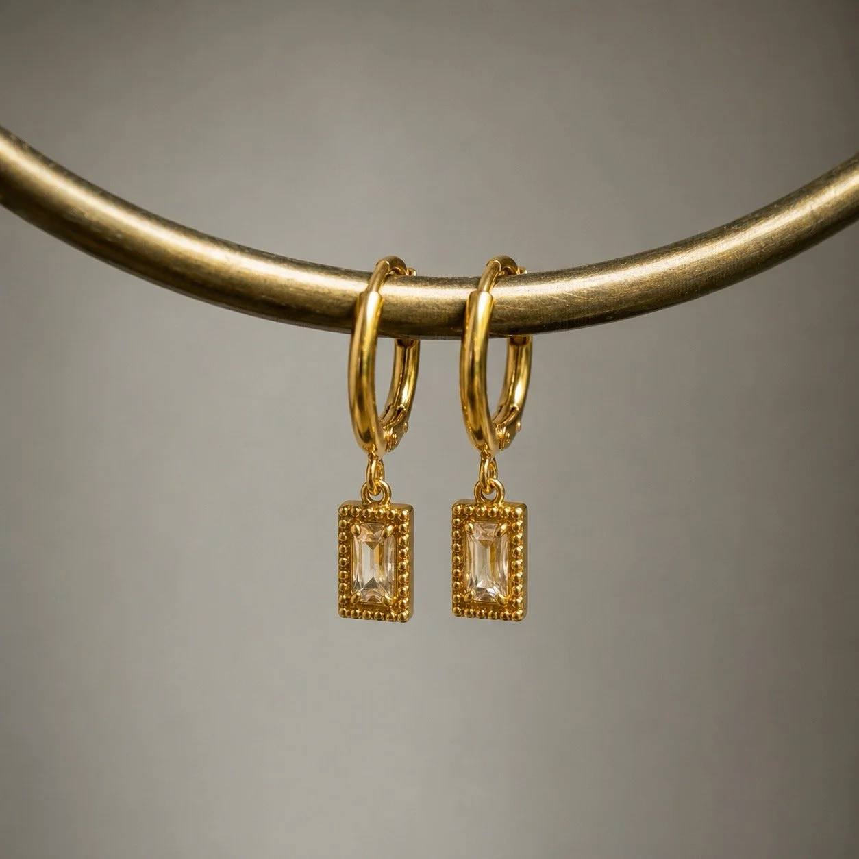 House Thirteen Glister Rectangle Huggie Earrings hanging on gold curved pipe display, cubic zirconia drop huggie earrings in stainless steel. Comfortable for everyday wear