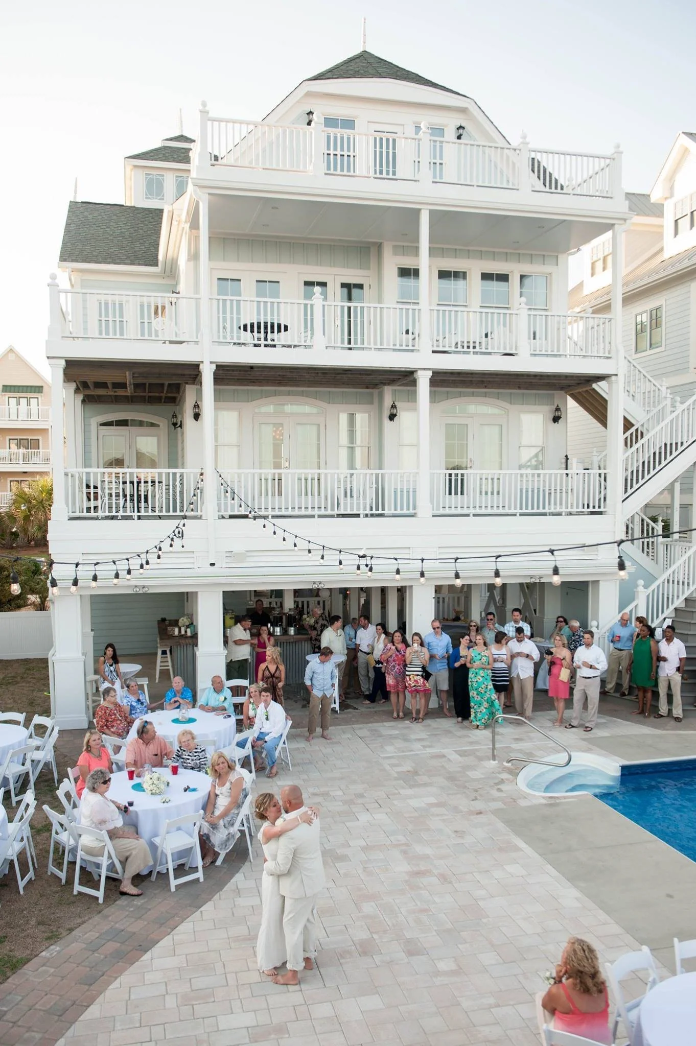People at a wedding reception celebrating outdoors near a large white house with multiple balconies and string lights. A couple is dancing in the foreground, and guests are gathered around tables and along the house's patio.
