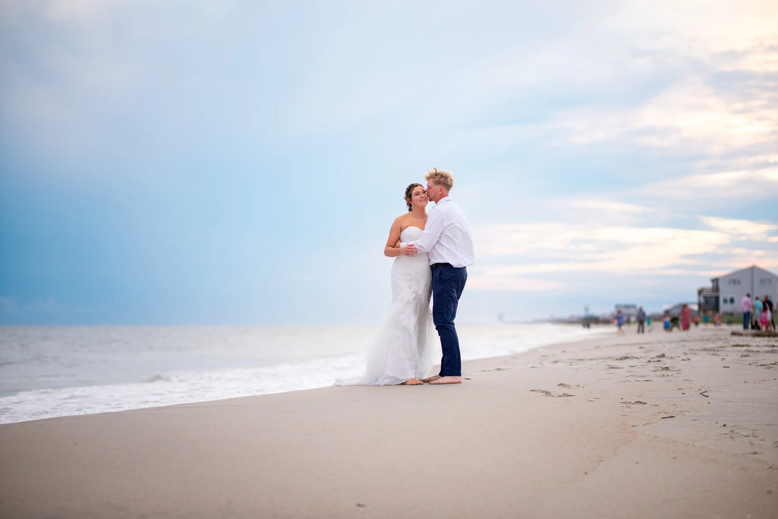 A bride and groom stand on a sandy beach, facing each other and smiling, with the ocean and a cloudy sky in the background during sunset or early evening.