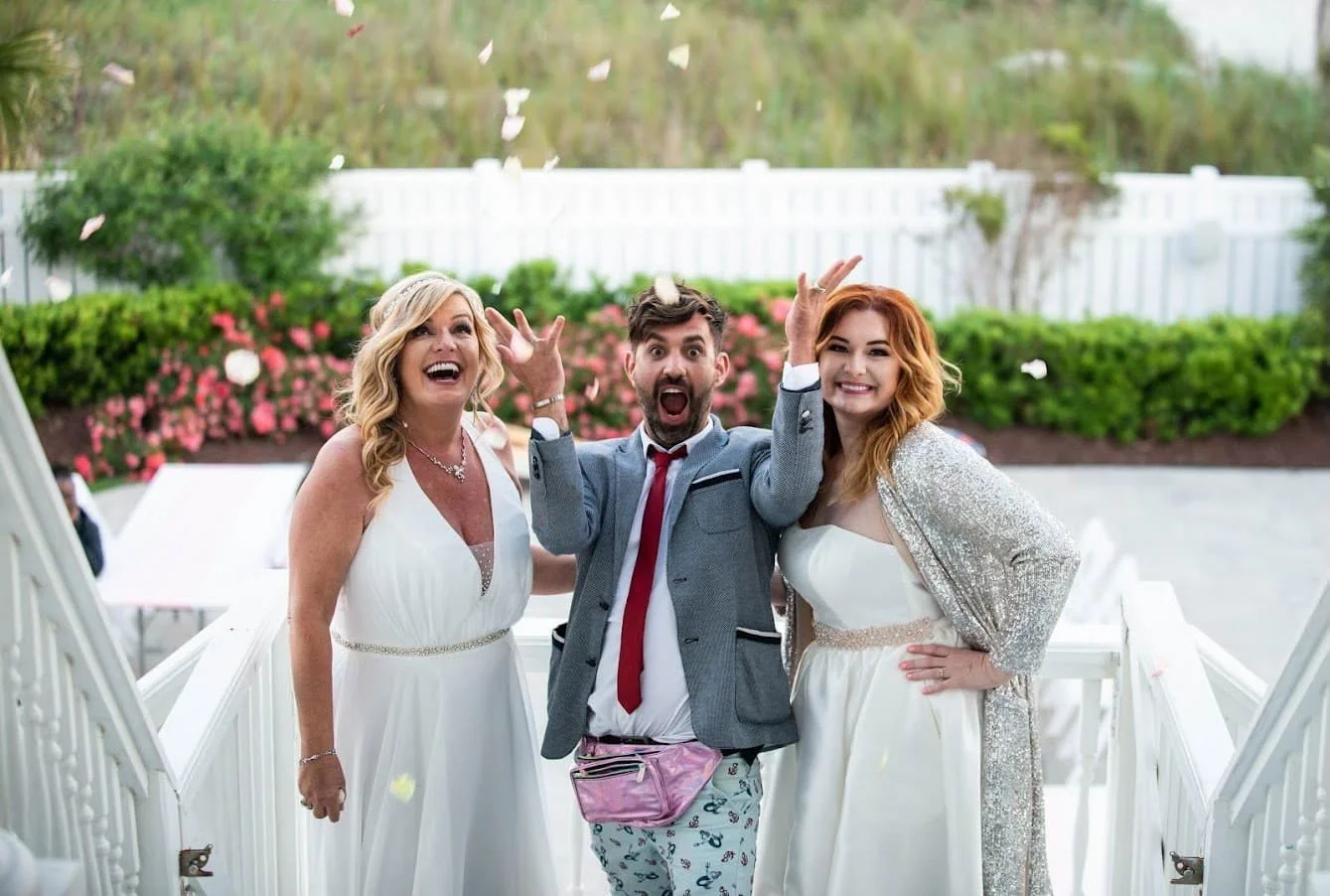 Three people at a celebration, throwing flower petals. Two women in white dresses and a man in a suit and colorful pants, smiling and celebrating outdoors.