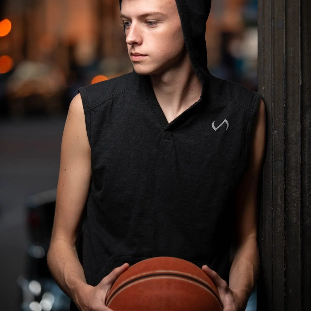 A young man wearing a sleeveless black hoodie with a hood and holding a basketball, standing near a wooden wall at dusk with blurred city lights in the background.