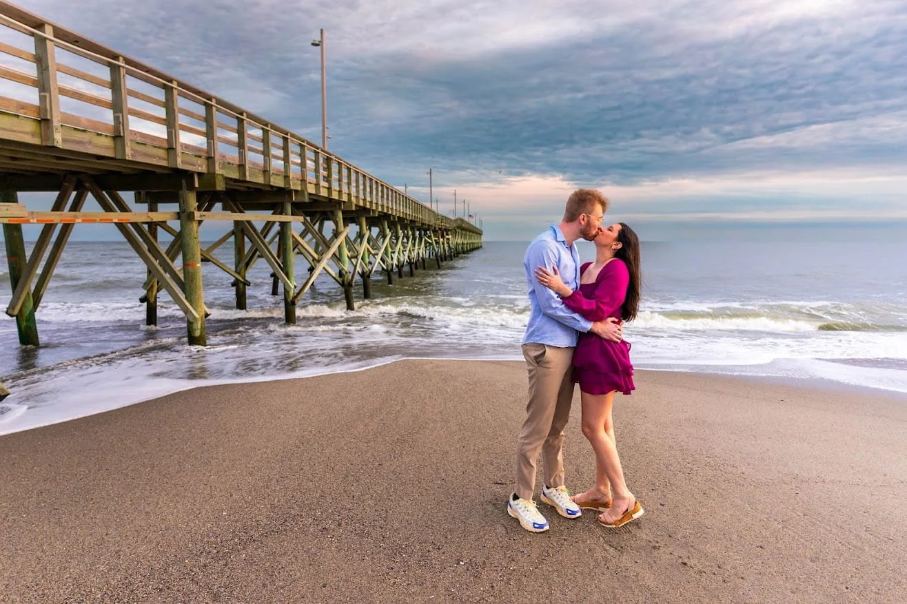 A couple kissing on a sandy beach near a wooden pier with the ocean and cloudy sky in the background.