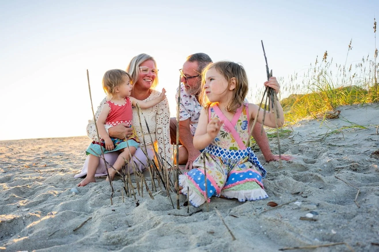 Family of four building a small fire on the beach at sunset, with sand and grasses in the background.